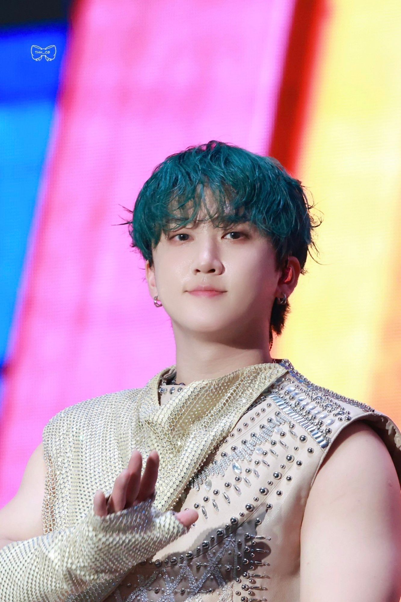 Changbin is wearing a beige sleeveless top with lots of silver studs and a beige blanket and gloves of the same fabric with lots of rhinestones. His hair is green. He is looking directly at the camera and is waving with his right hand.