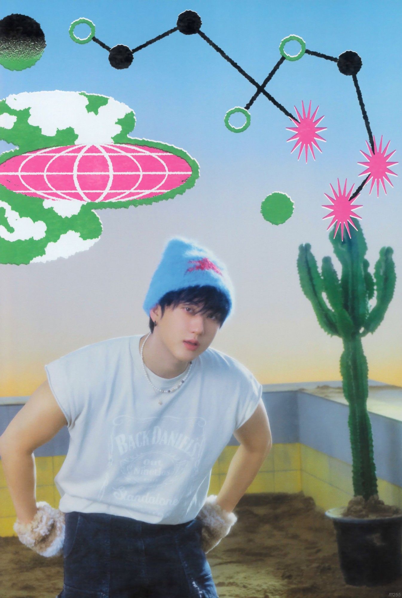 Changbin is dressed in a white t-shirt and dark jeans, white plush gloves and a blue wool hat with a red star in the center. He is standing with both hands in his pockets. There are cacti in the picture. He's leaning forward a little. He is in an empty pool with sand and yellow tiles.  