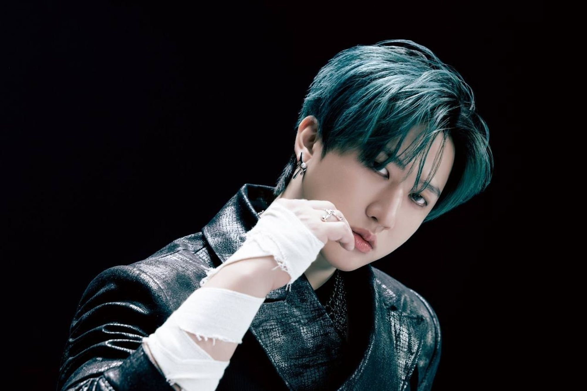 Changbin is dressed in a black leather suit and his right hand is raised in front of his face and his fist is clenched. His forearm and hand are bandaged with white fabric. His head is tilted to the left. The background is completely black.