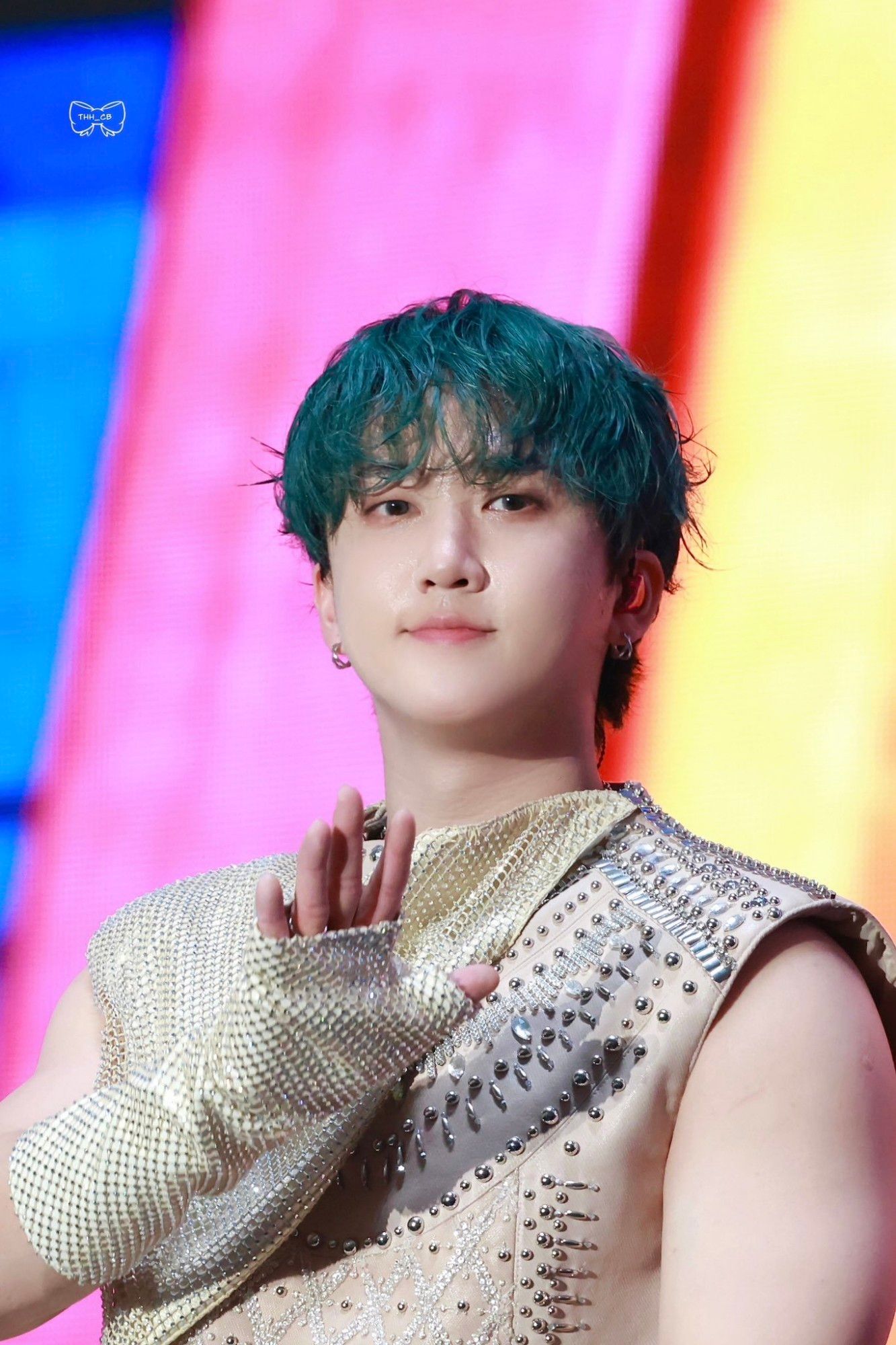 Changbin is wearing a beige sleeveless top with lots of silver studs and a beige blanket and gloves of the same fabric with lots of rhinestones. His hair is green. He is looking directly at the camera and is waving with his right hand.