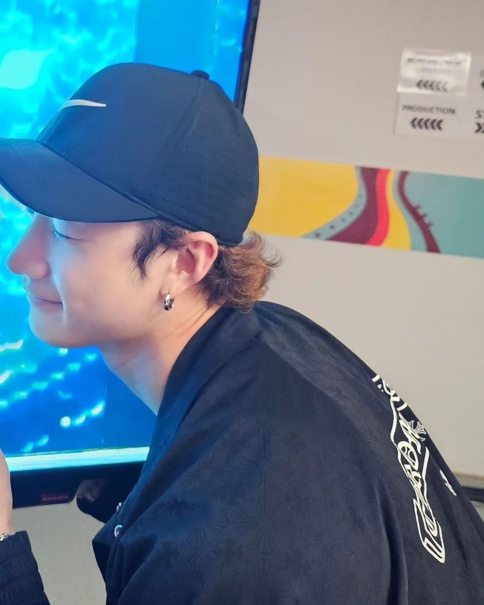 This is a picture of Bang Chan. He is wearing a black long-sleeved shirt with a white lettering on the back and a black Nike baseball cap. It is a profile picture of him and his body is turned to the right. He has a small smile on his face and his eyes are closed.