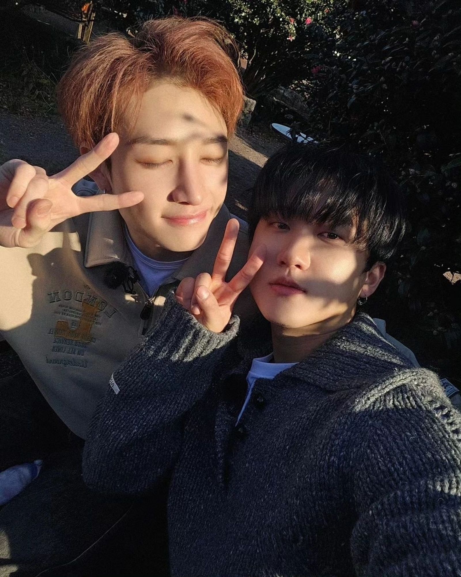 This is a selfie of Changbin with Bang Chan. Changbin is wearing a gray sweater and Bang Chan is wearing a beige hoodie. The angle of the photo is from above. Changbin is taking the photo. He is looking at the camera and is making the peace sign with the fingers of his right hand near his right cheek. Bang Chan is facing Changbin (on his left) with his eyes closed and is also making the peace sign with his fingers of his right hand near his right eye. In the background there are trees