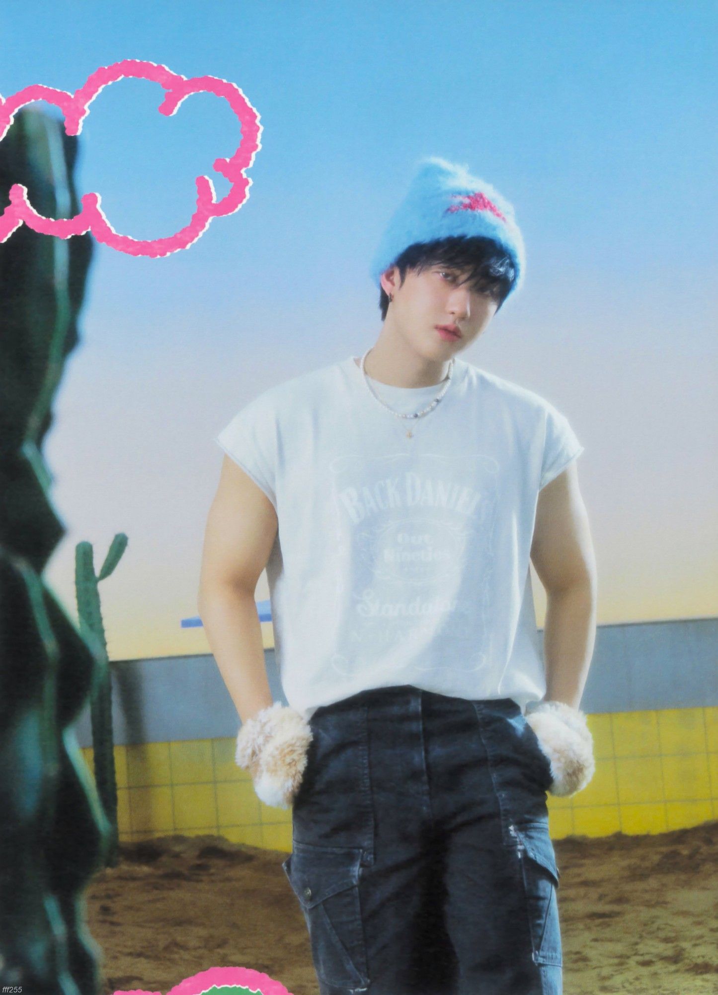 Changbin is dressed in a white t-shirt and dark jeans, white plush gloves and a blue wool hat with a red star in the center. He is standing with both hands in his pockets. There are cacti in the picture. He is in an empty pool with sand and yellow tiles. 