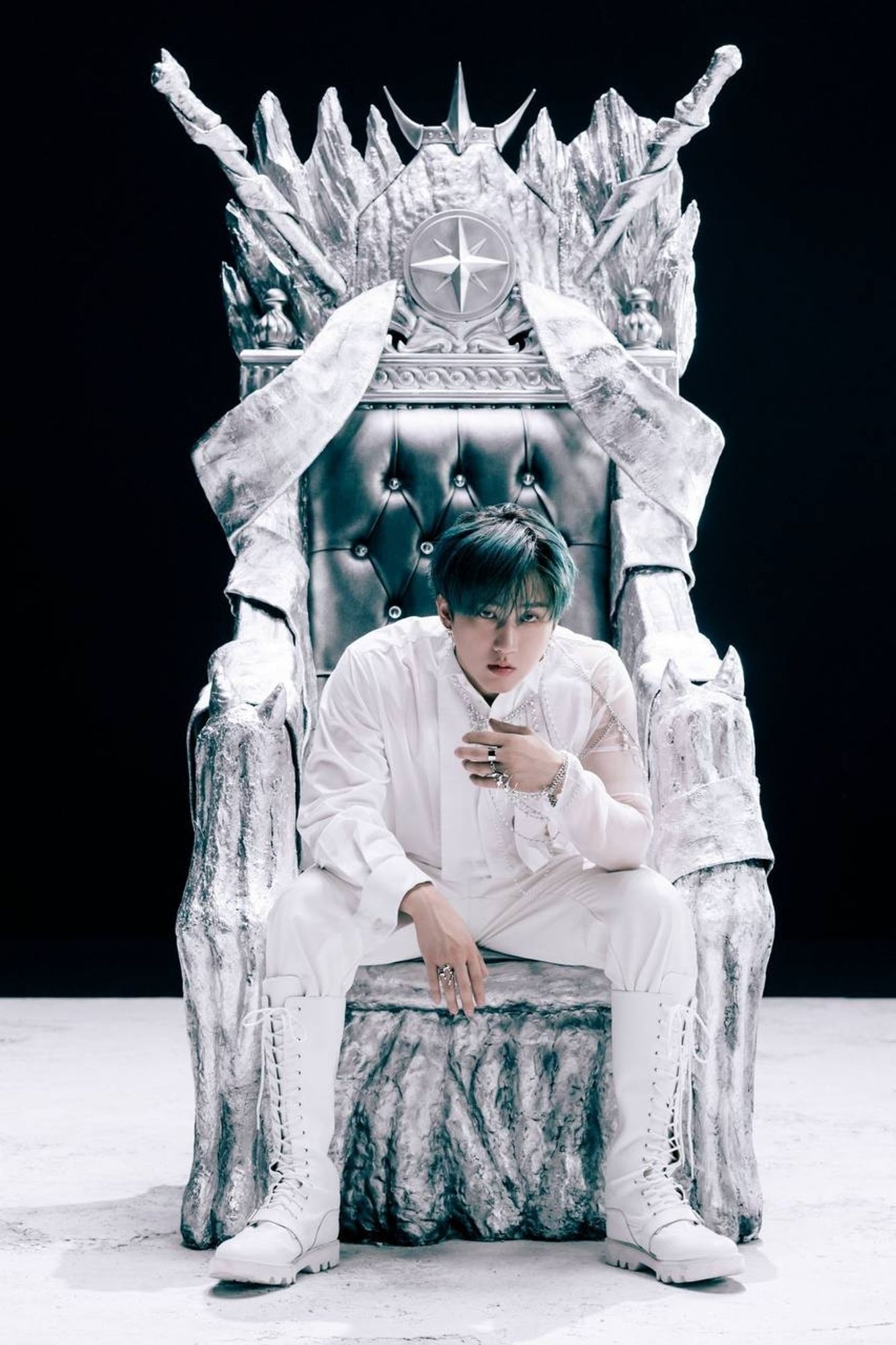 Changbin is wearing a white long-sleeved blouse made of a semi-sheer fabric, white pants, and white knee-high boots. He is sitting on an imposing silver throne, and on top of the throne is a compass (the Stray Kids symbol). He is leaning forward, his legs are spread, and his right arm is resting on his right thigh and his left elbow is resting on his left thigh. His left hand is covered in silver rings and is on his chest, and his right hand is hanging down. He is facing the camera. The background is completely black and the floor is white.