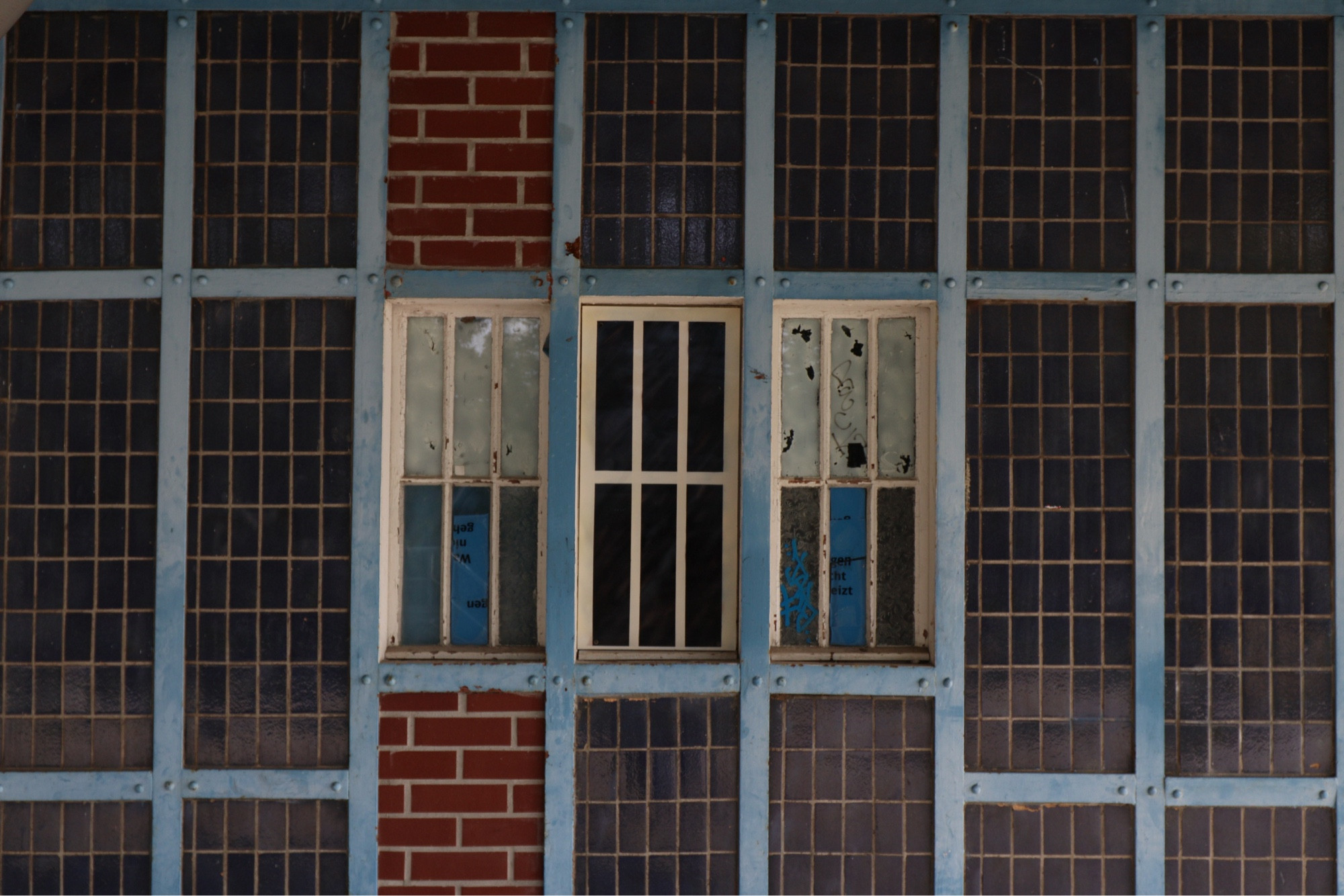 Broken windows of a disused platform facility, blue ironwork makes a grid over dark slate tiles. A single column of red brick aligns with the leftmost of three windows