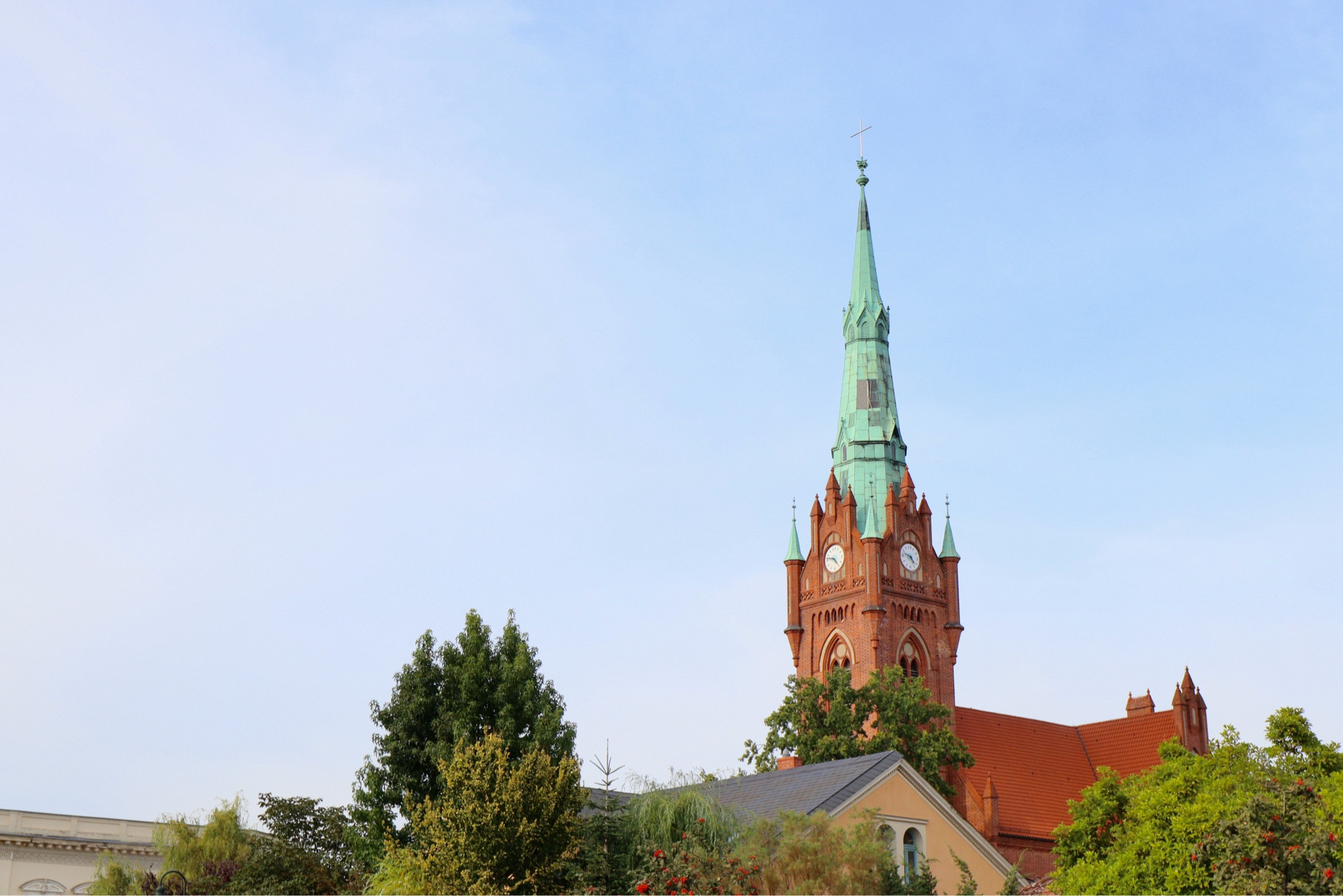 Pfarrkirche Herz Jesu, a red brick spire with a green copper steeple raises above the roofs and trees into a hazy blue sky