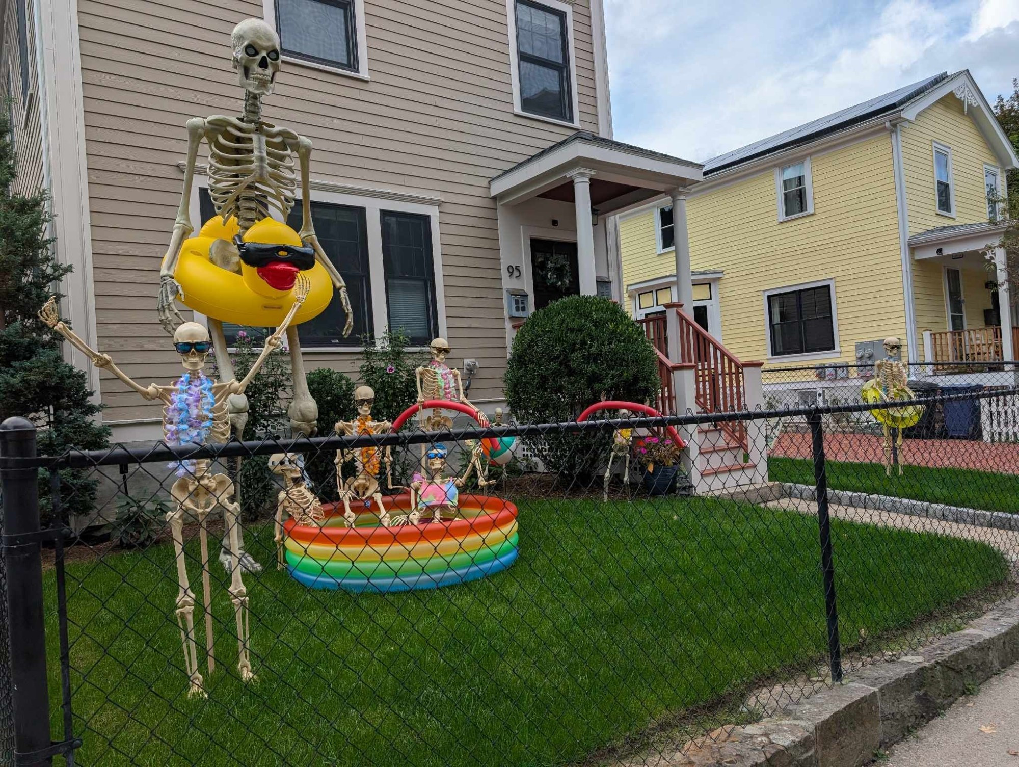 a giant skeleton with a yellow duck pool floaty and numerous smaller skeletons wearing leis and chilling in a kiddie pool with swim floats and pool noodles