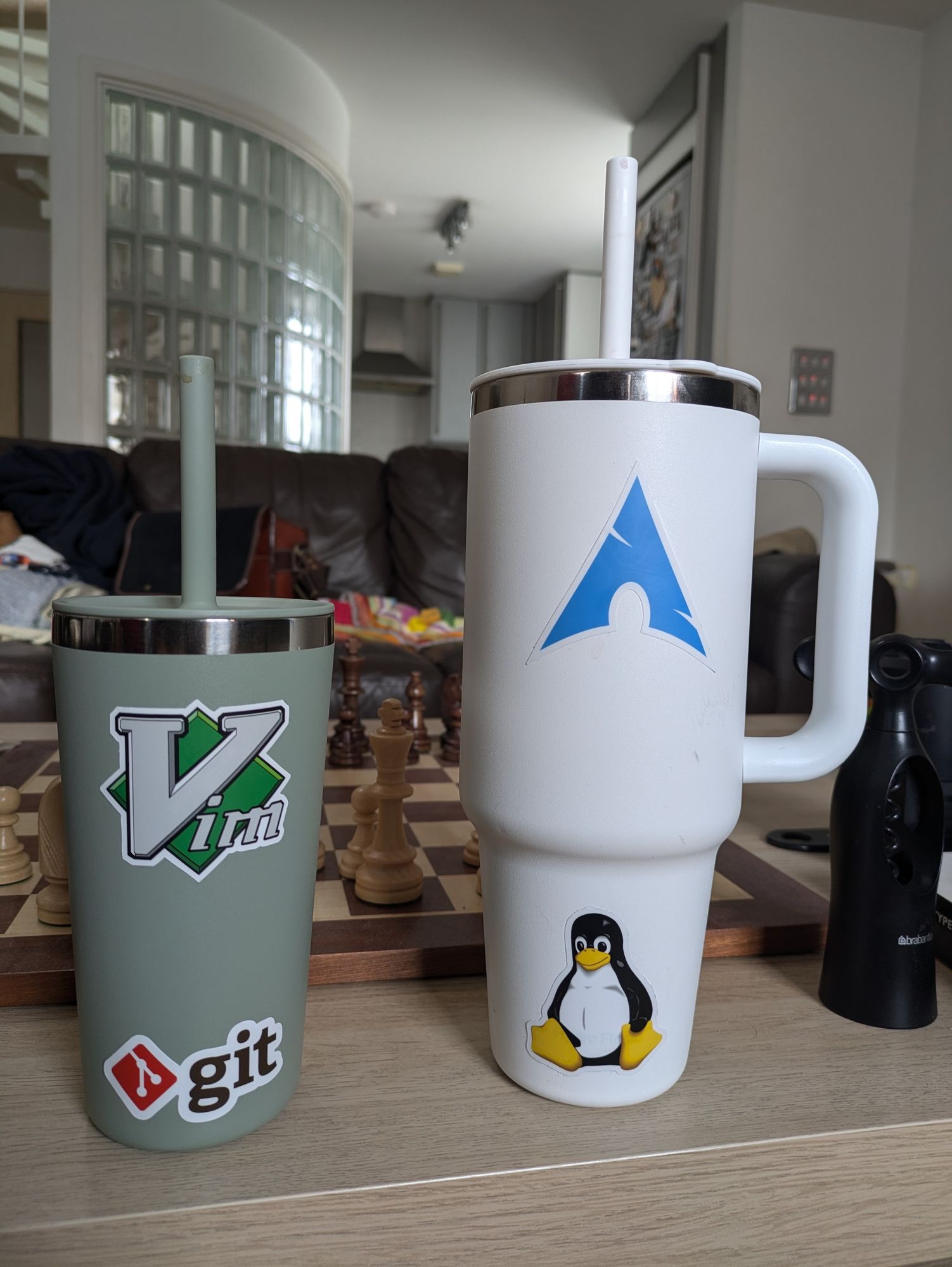 A picture of two travel mugs, one small and green, one big and white with a handle.  Both are on my coffee table, both have straws.  Near them is a corkscrew.  Behind them is a large chess board, my brown leather couch, and the spiral staircase to my bedroom behind glass panelling.

Each of the travel mugs has had their logos covered up by stickers: the green one with the vim and git logos, the white one with the Linux penguin and the Arch Linux logo.