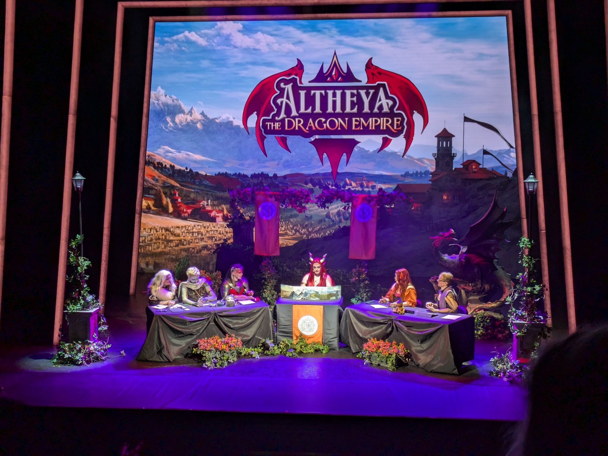 A photograph of a theatre stage on which five players and one DM are playing Dungeons and Dragons.  They are the High Rollers.  A backdrop depicts a vast countryside landscape with the title, "Altheya, the Dragon Empire," above it.

The players are dressed as their characters, with one woman wearing white hair with a white fur top, one man with grey skin and white hair with what looks like leather armour and a scarf across his neck, another woman wearing plated armour over red clothes and with red face paint on the upper half of her face, another woman dressed like a normal peasant, and another man wearing wizard robes.  The DM appears to be a man wearing a red dress with long, bright red hair, and horns protruding from his head.