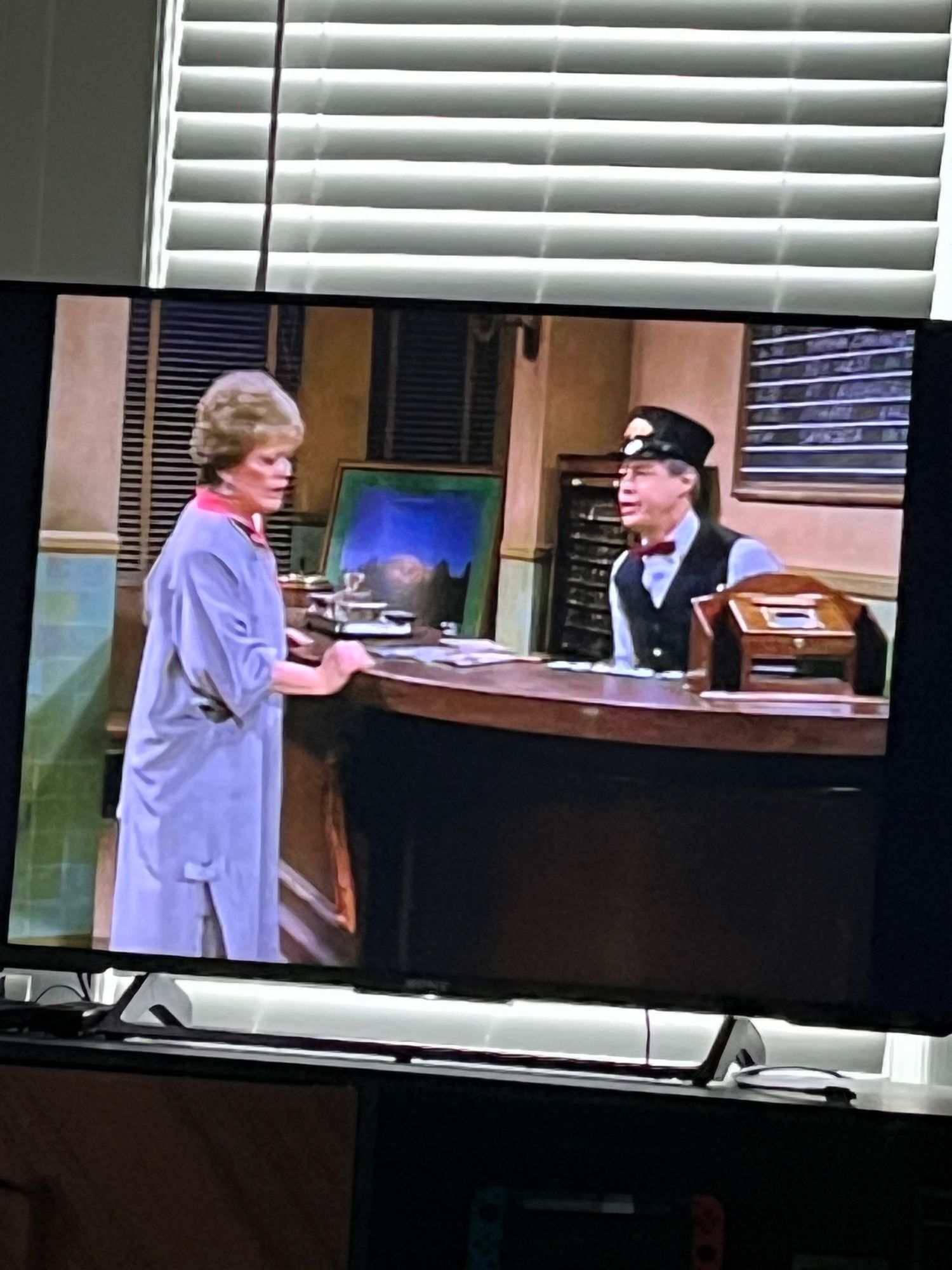 Photo of a tv showing Blanche of Golden Girls (long purple coat and grayish brown hair) talking to a train ticket guy at a desk.