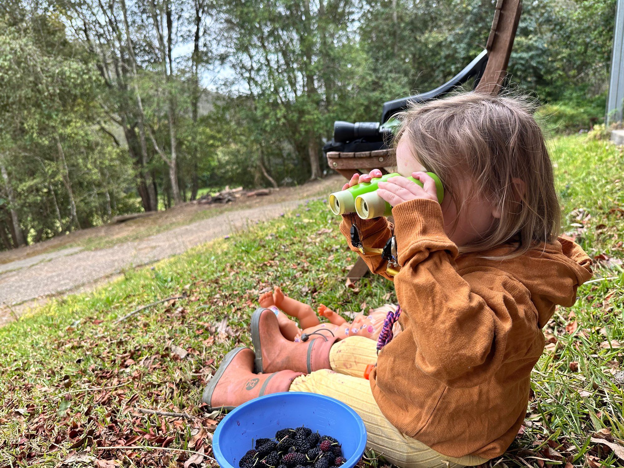 A toddler in a brown hoodie, yellow pants and pink boots, sitting on the grass using a pair of small green binoculars backwards.