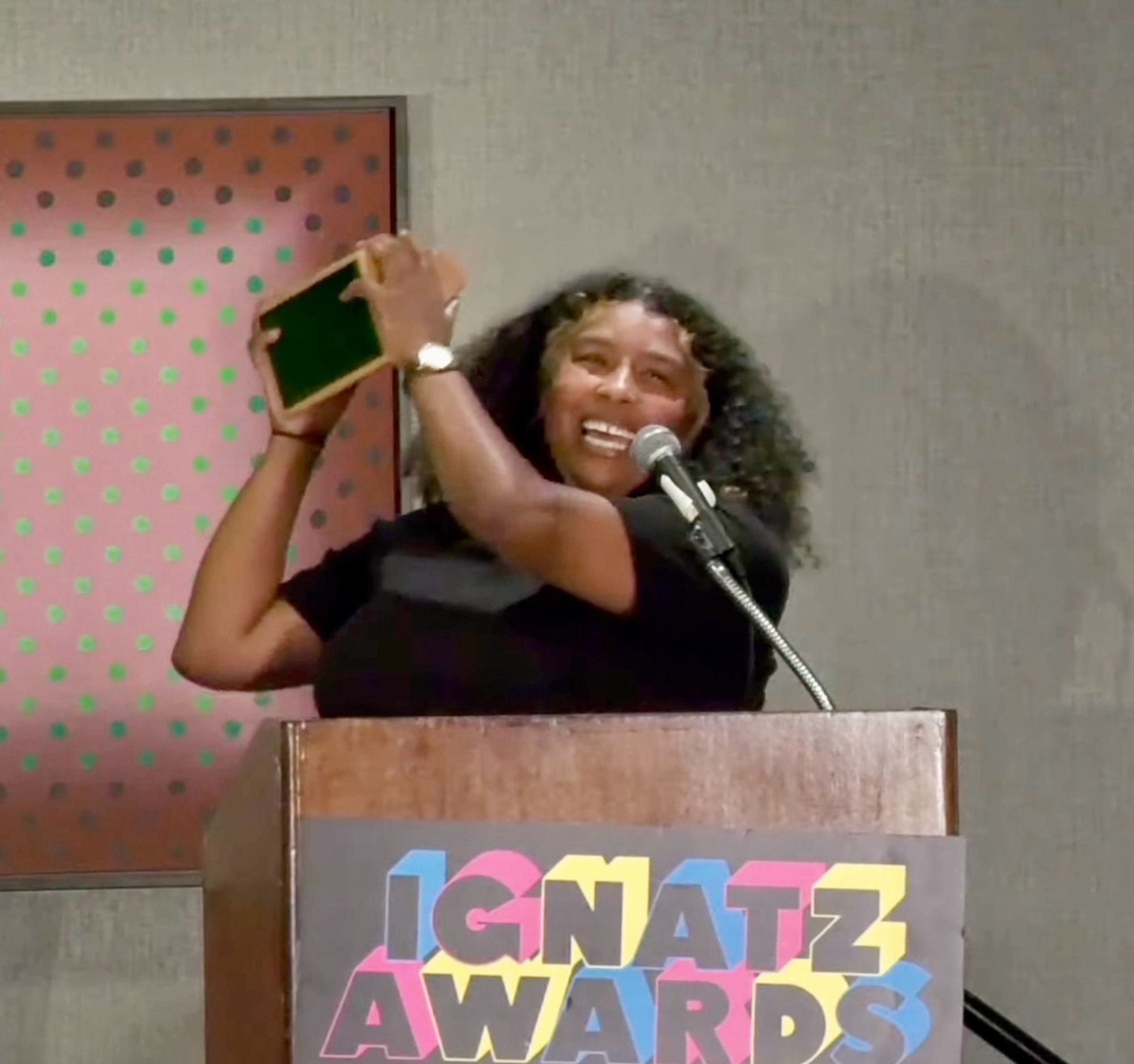 Black girl smiling and holding up an award in front of a podium.