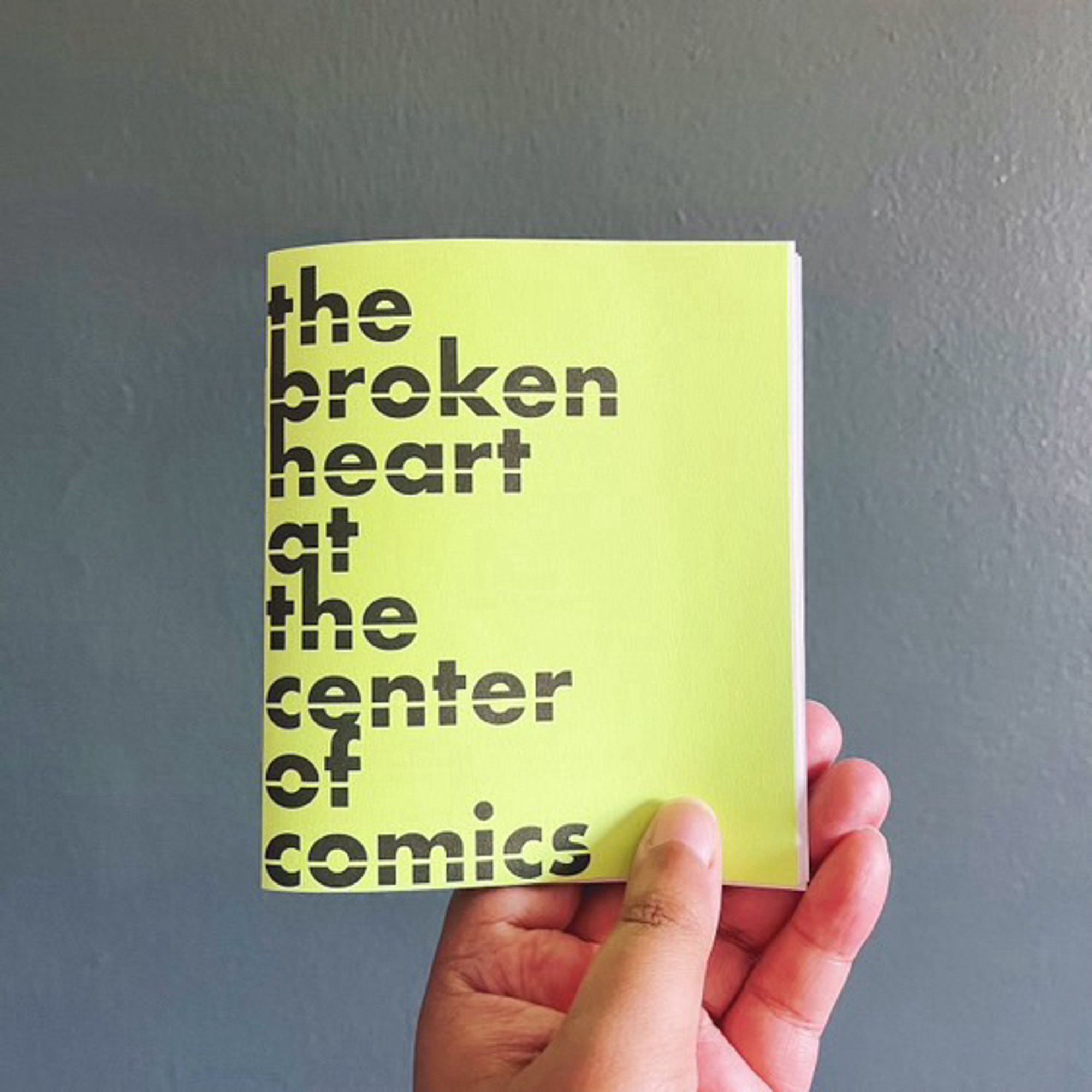 A hand holding a lime green zine titled “the broken heart at the center of comics”