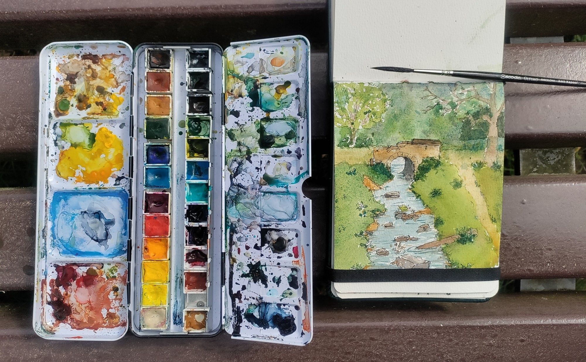 Watercolor set and sketchbook with a landscape sketch of creek and bridge