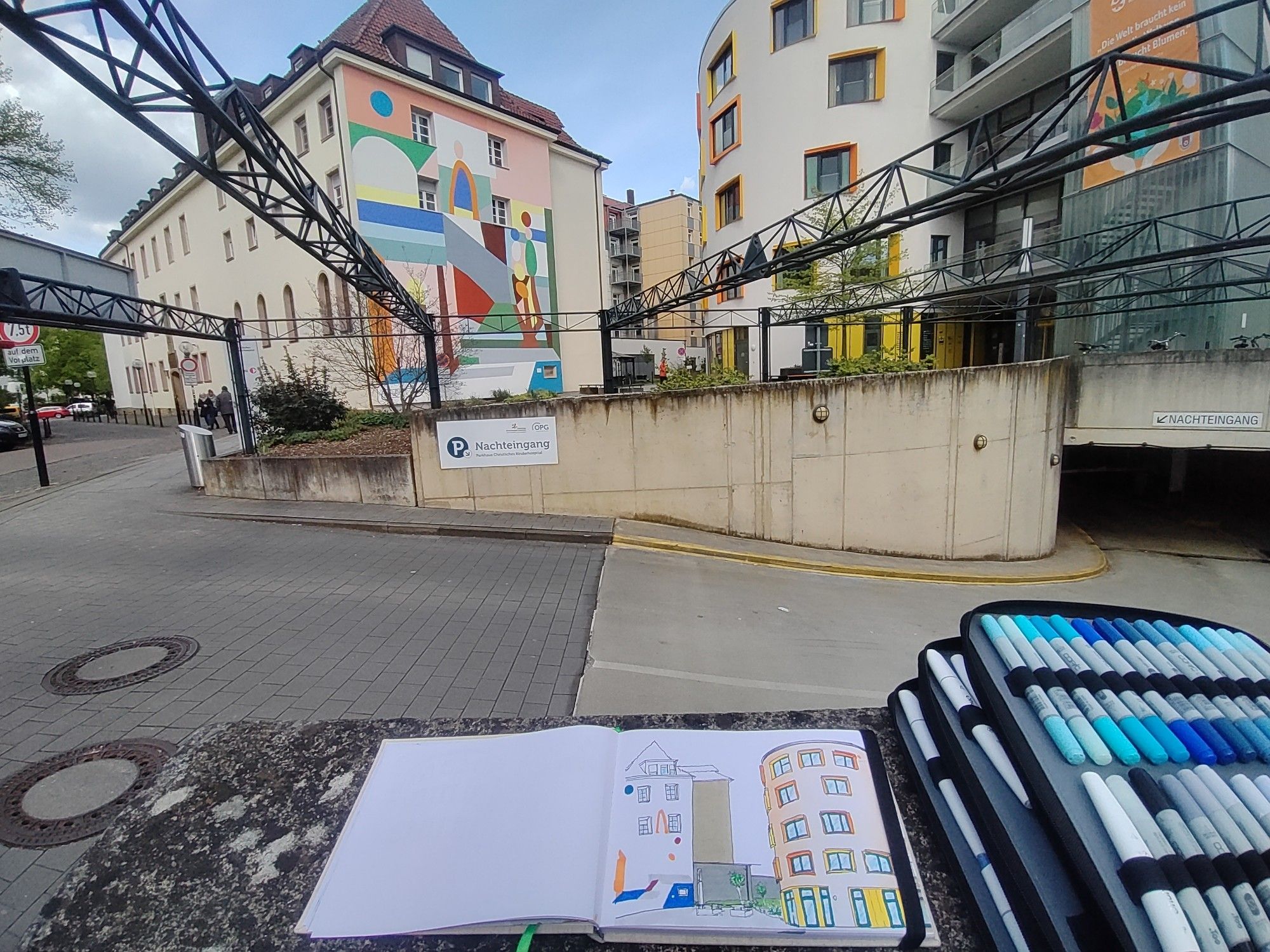 Wip Urban sketch with markers shown next to it, of the colorful buildings of a childrens hospital, seen in front of the real buildings