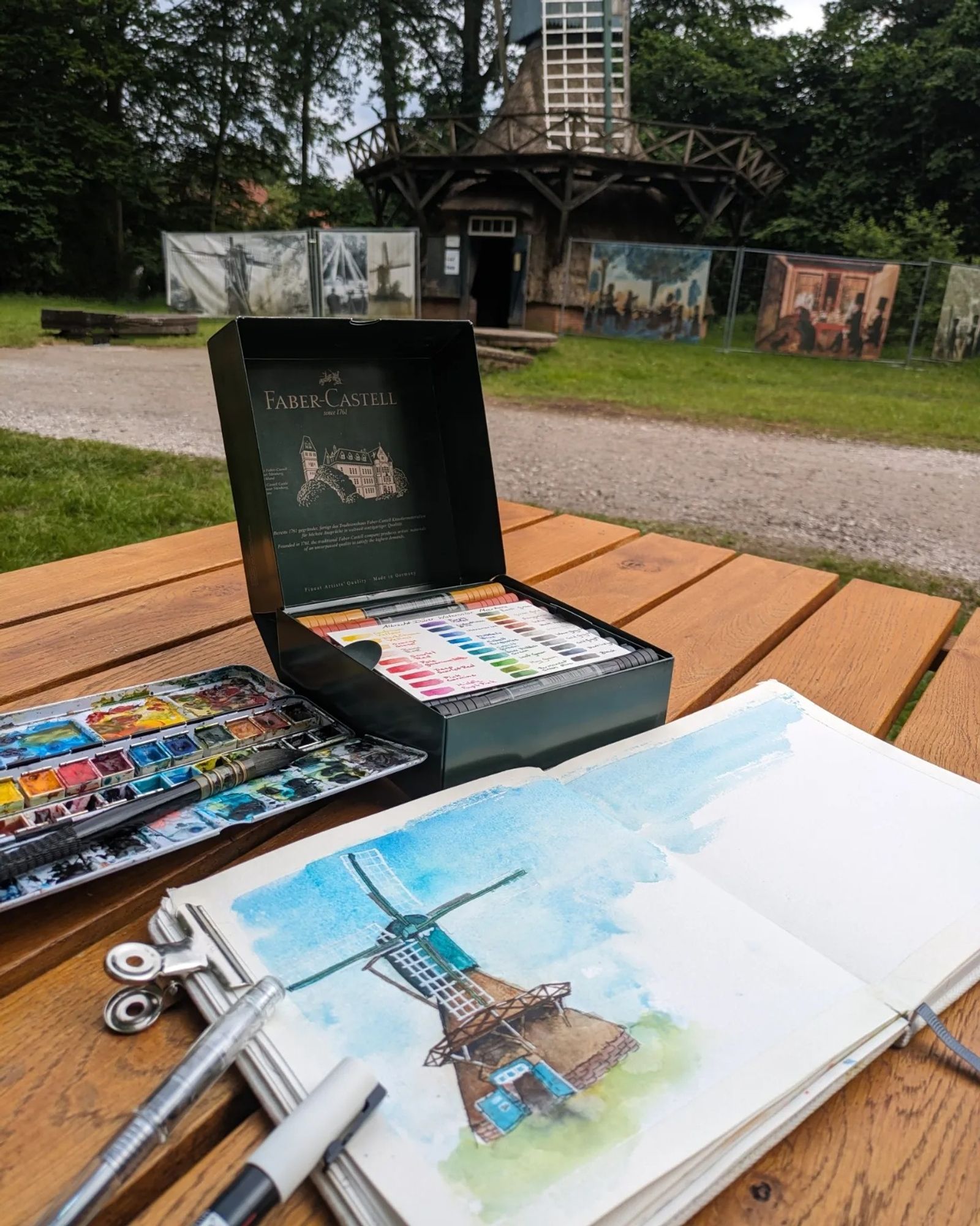 A windmill watercolor urban sketch with a brown base and bluegreen top with the mill in the background. The mill is standing in the outdoor museum of historical farm buildings in Cloppenburg, Germany