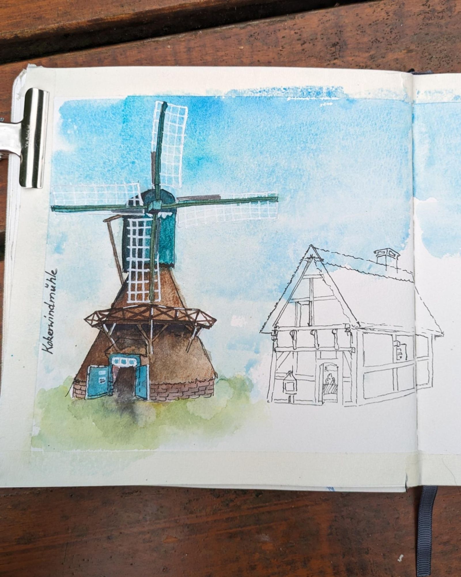 Sketchbook-page: A windmill watercolor urban sketch with a brown base and bluegreen top. The mill is standing in the outdoor museum of historical farm buildings in Cloppenburg, Germany