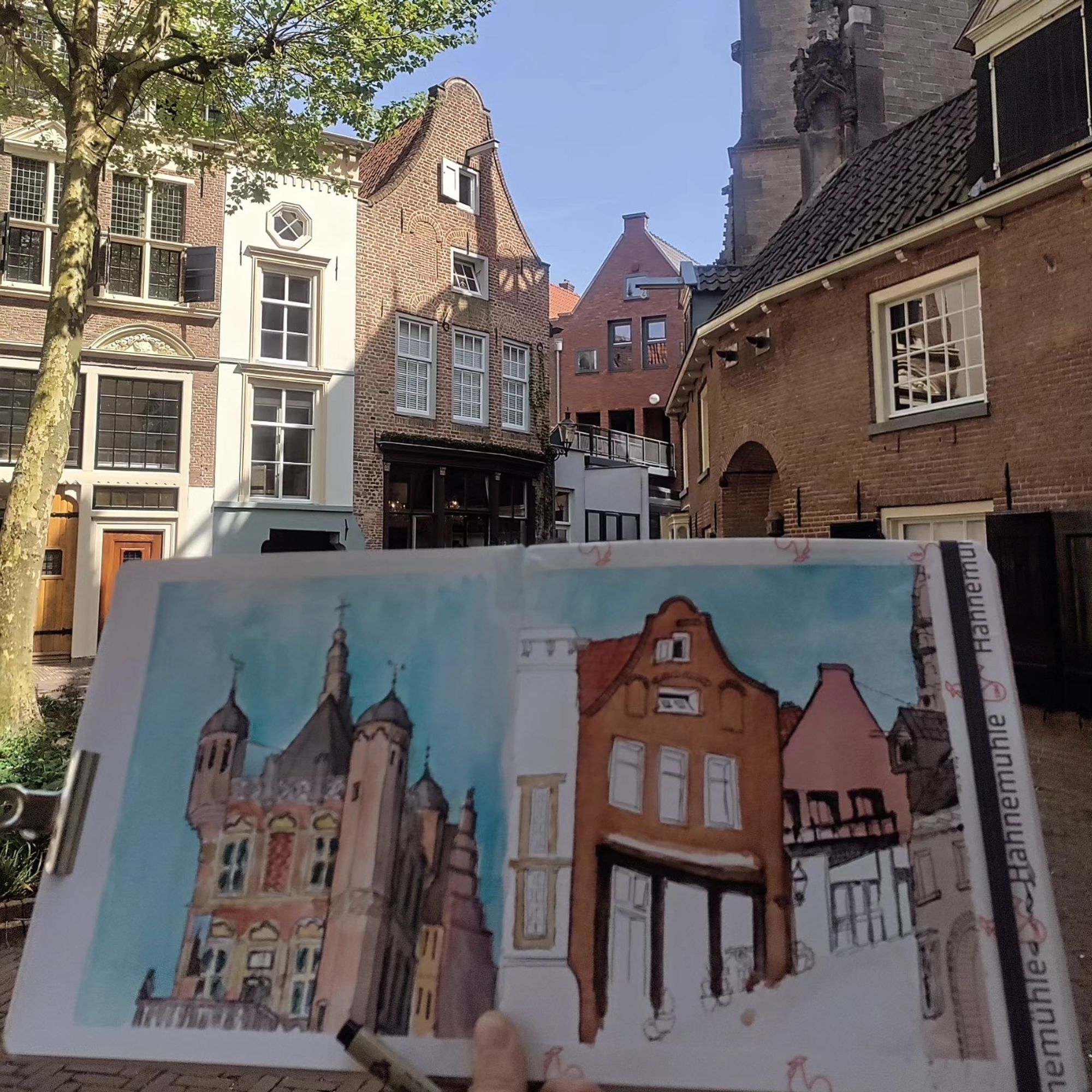 Sketchbook with a sketch of a square behind a church in Deventer, NL held in front of the real thing