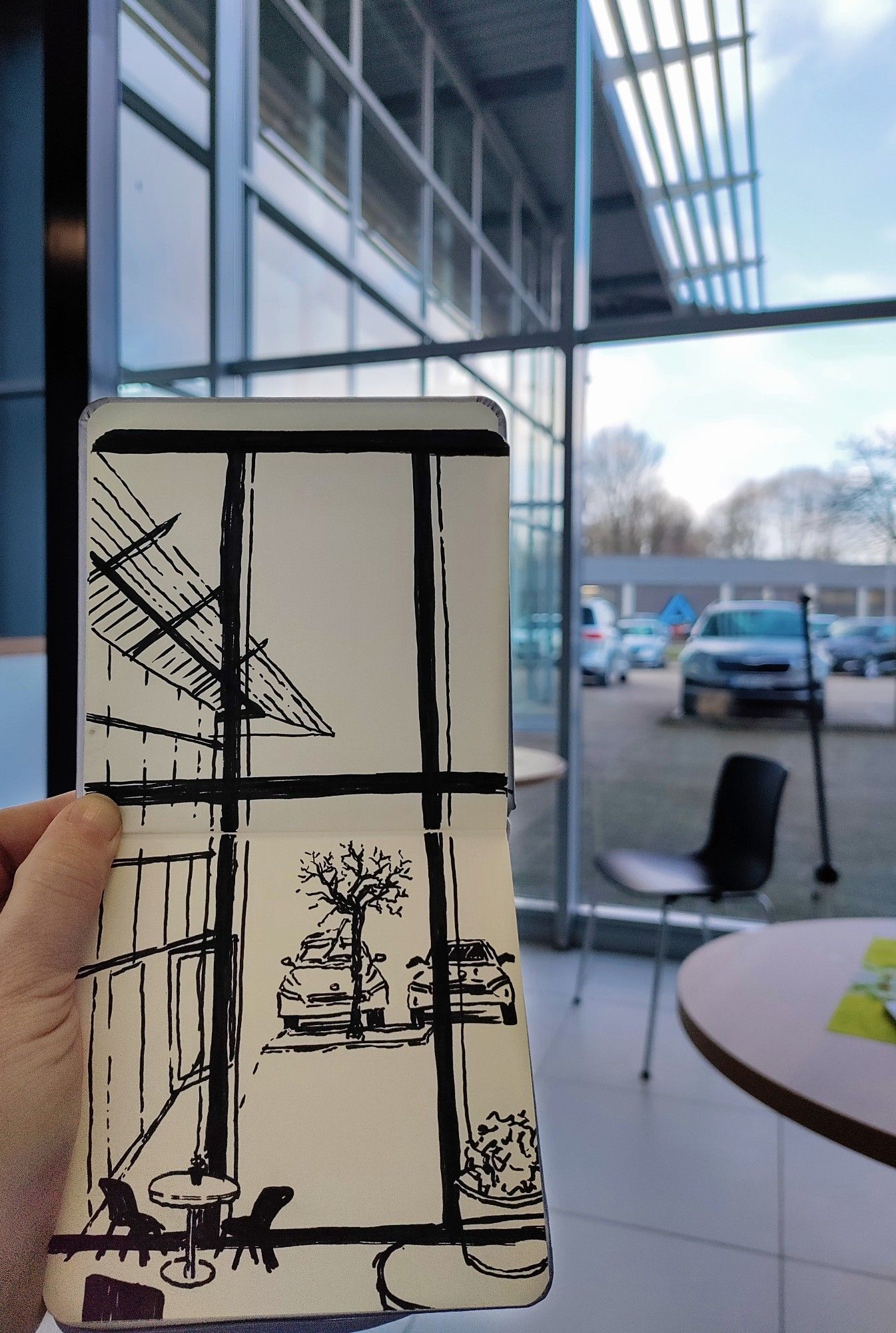 A sketchbook held open with the view sketched from a car dealership waiting area through the windows