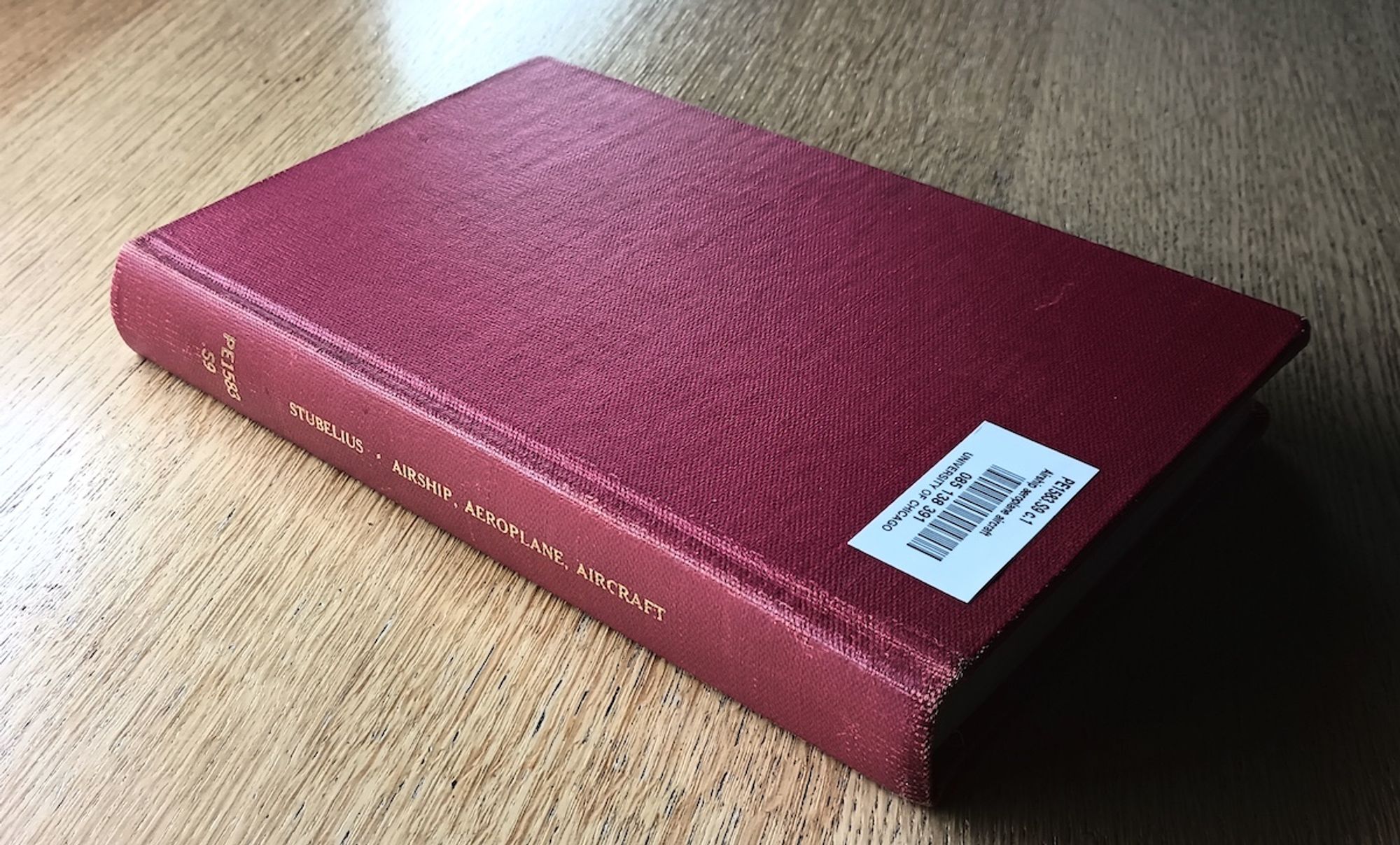 Photo of a red book on a wooden tabletop. The cover is plain with no pictures; text on the spine is stamped in gold letters: "Stubelius - Airship, Aeroplane, Aircraft" along with a catalogue number.  A modern barcode sticker attached to the front cover identifies this as  belonging to the University of Chicago Library.  Photo copyright 2024 by Willam S. Higgins.