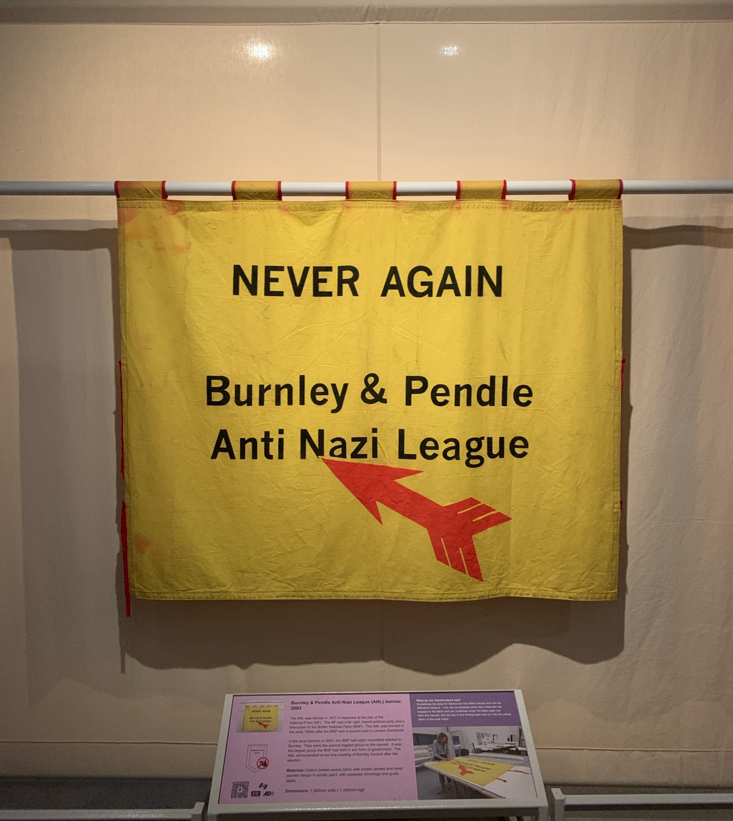 A yellow hanging banner saying “NEVER AGAIN   Burnley & Pendle Anti Nazi League” with an arrow underneath it.