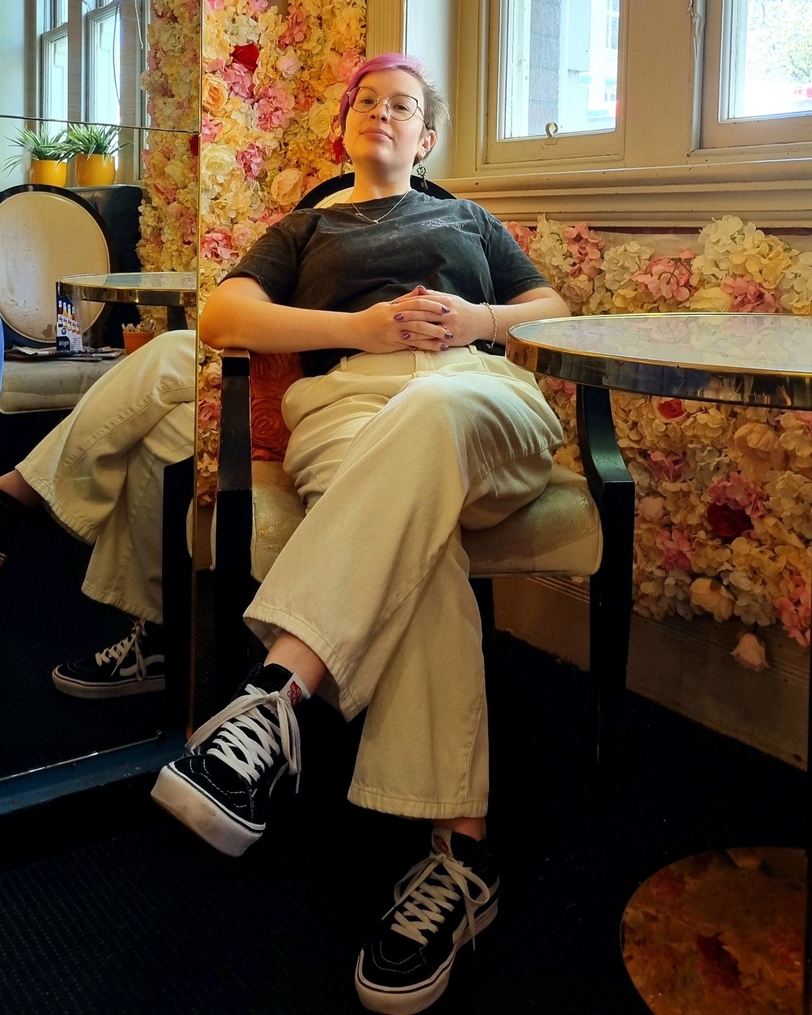 a low angle photo of Louise sitting at a small table and chairs with flowers all over the floor behind her, she's trying to have a stern expression but it isn't quite working