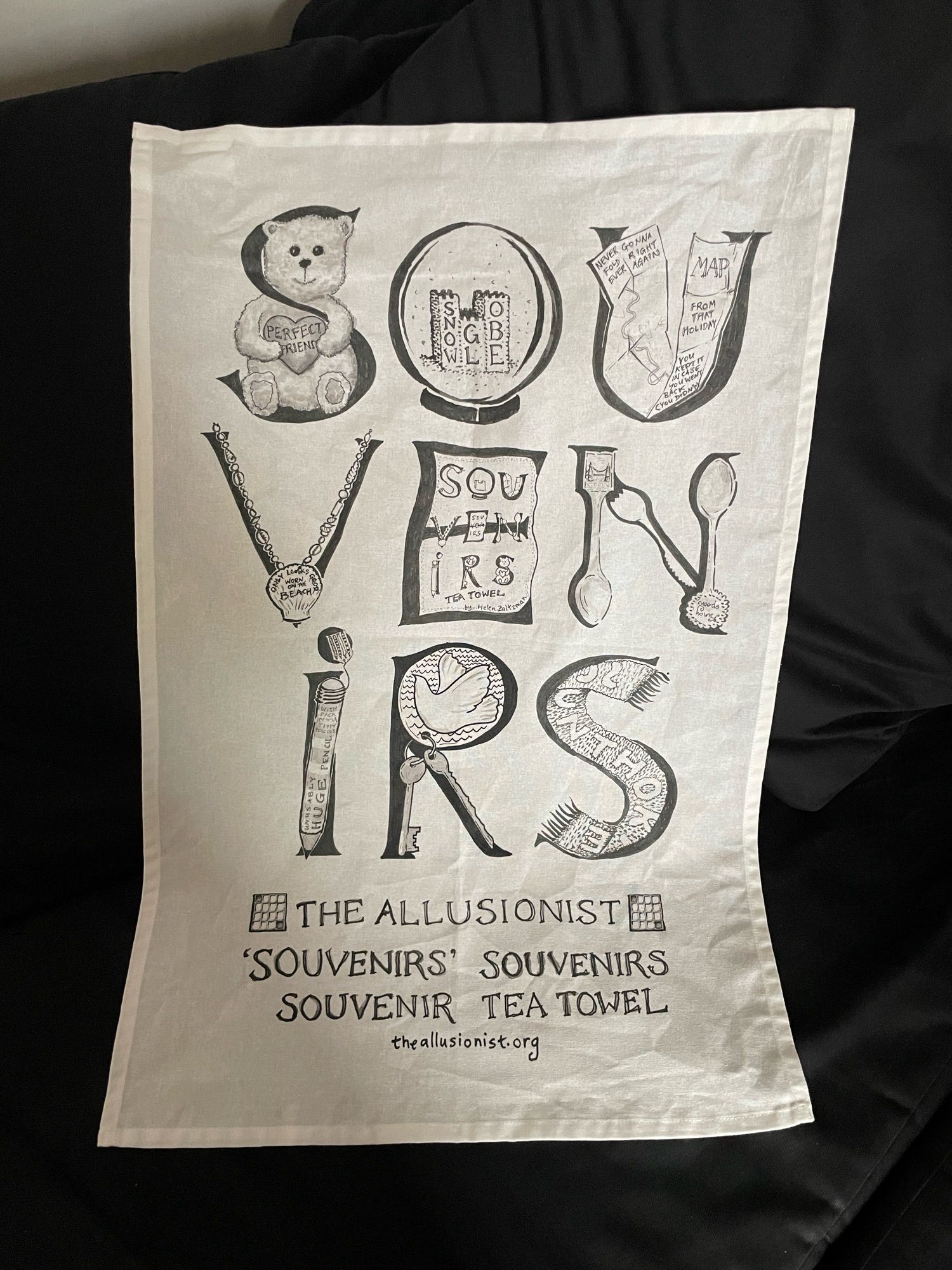 A tea towel that says Souvenirs on it and each letter of the word souvenirs has a hand drawing of a different souvenir item: teddy bear, keyring, snow globe, scarf, pencils, spoons, shell necklace, map