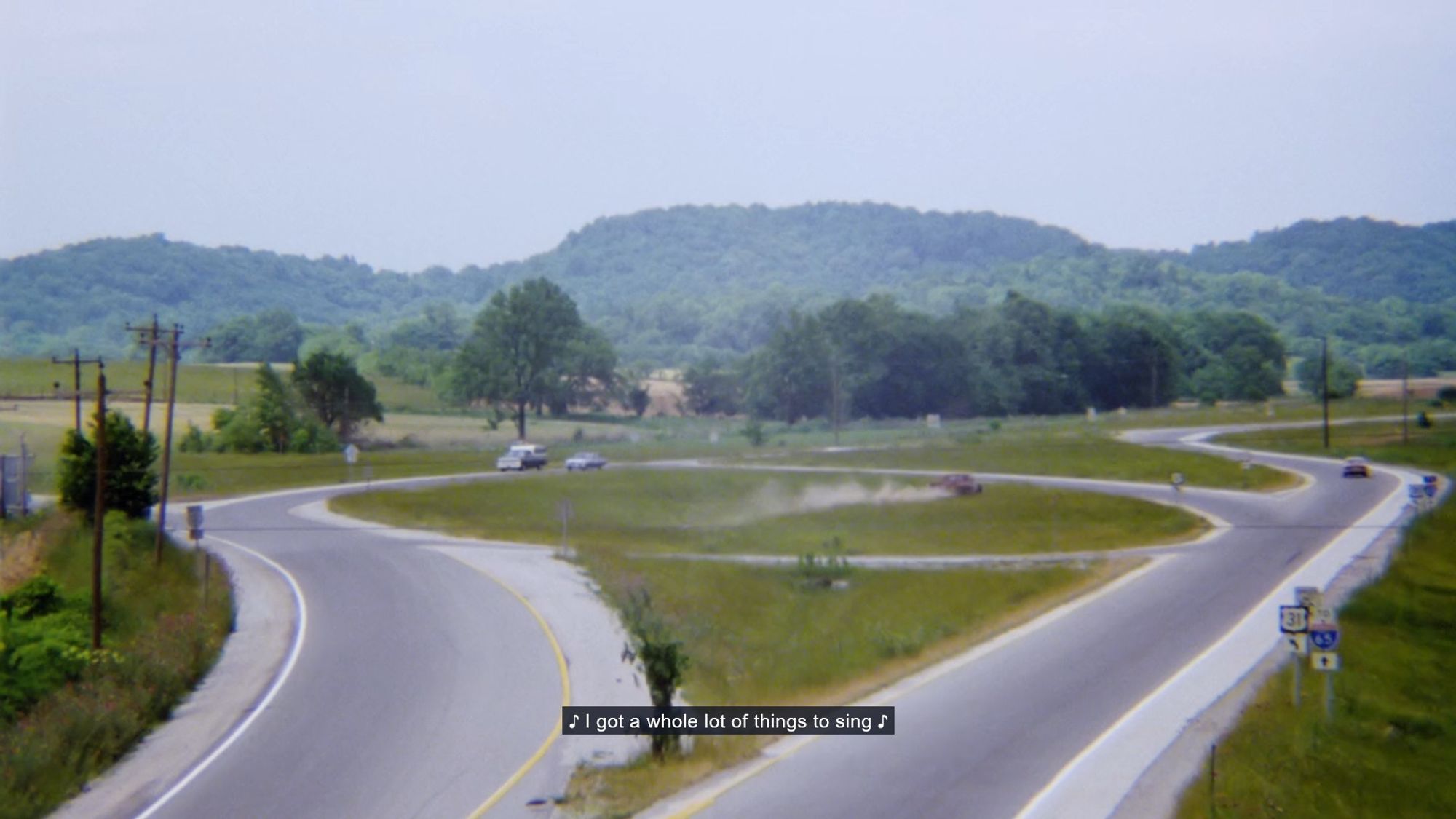 Film cap of a extra wide shot of a hot rod pickup truck screaming directly across the middle of a two large traffic circles, kicking up all kinds of trucker dust which other cars must eat, while the caption underneath features the lyrics of the current song playing on the soundtrack: "I got a whole lot of things to sing"