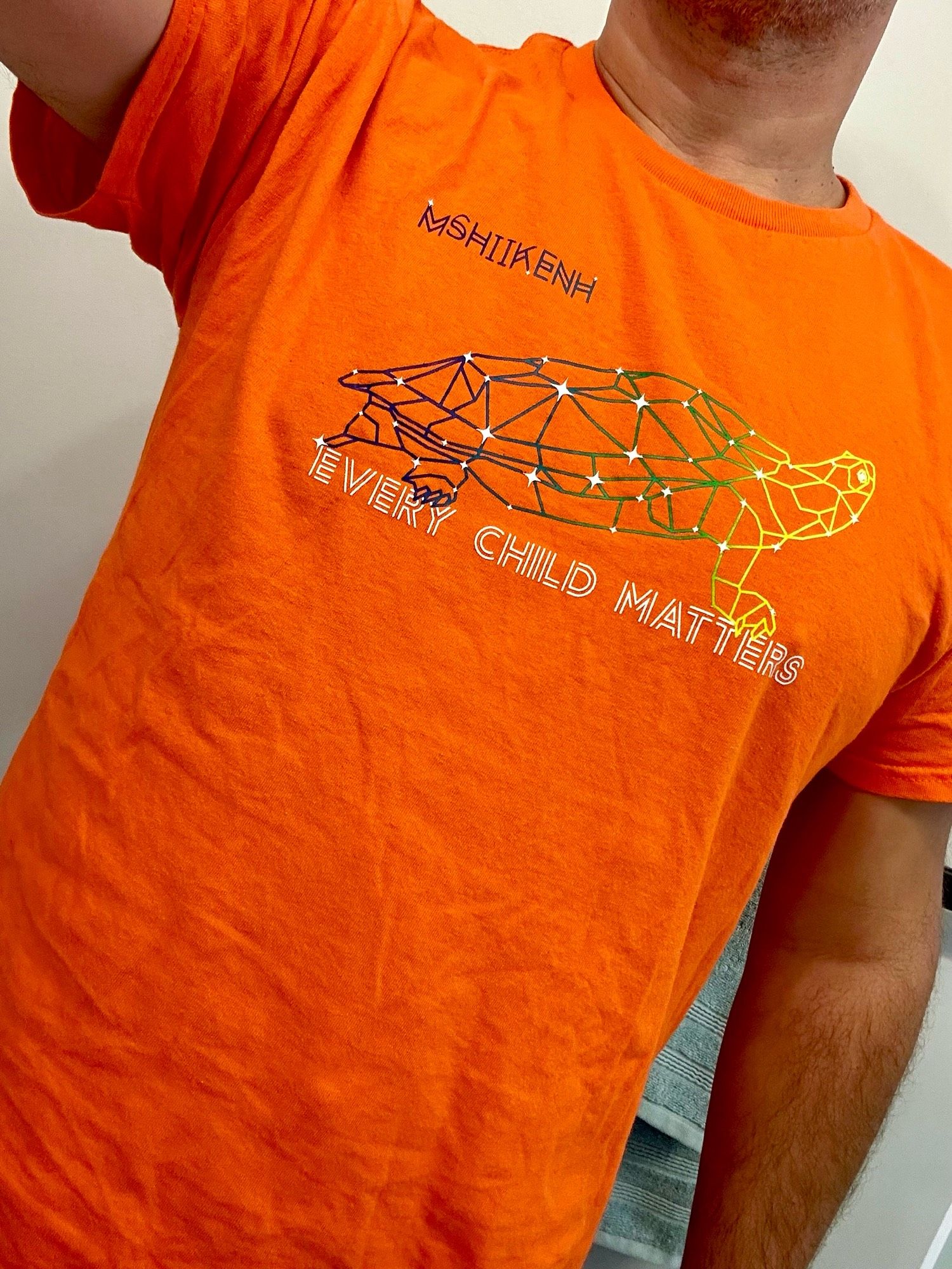 Closeup of orange t-shirt from Nish Tees. Vibrant orange, has a turtle constellation on it, with the words “Every Child Matters” and “mshiikenh” (Nishnaabemwib word for “snapping turtle”)