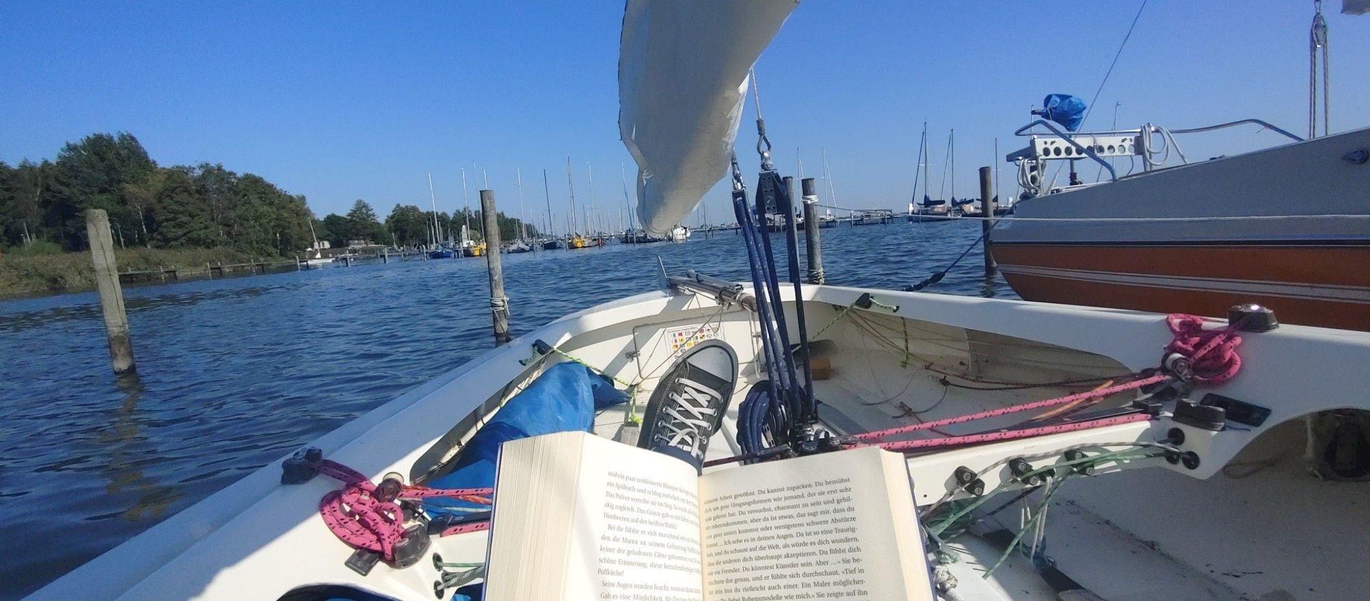 Blick über eine Segeljolle richtung Heck, im Hafen liegend. Im Vordergrund ein aufgeschlagenes Buch.