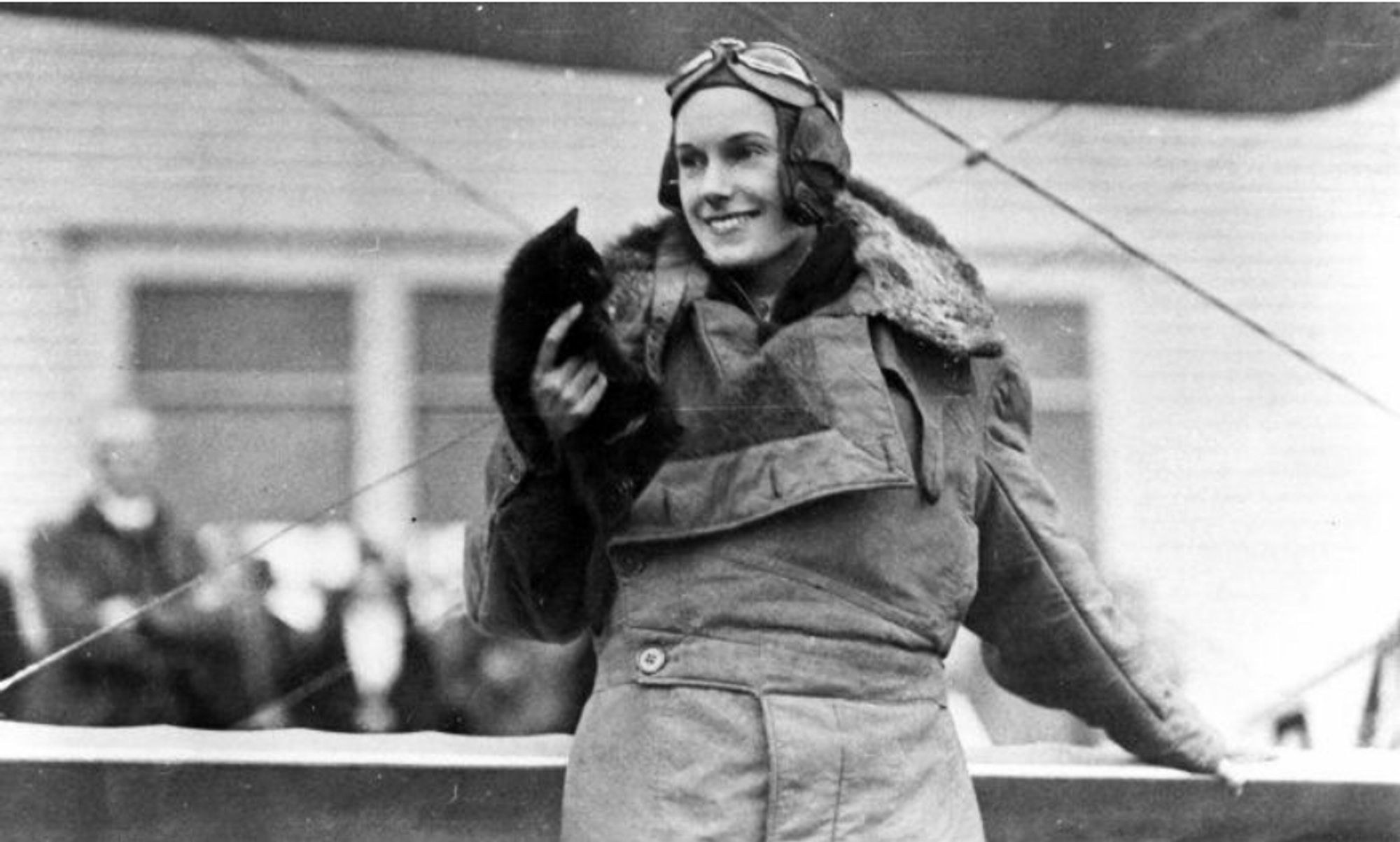 Jean Batten in her pilot's gear standing in front of her plane, holding a small black cat in one hand.