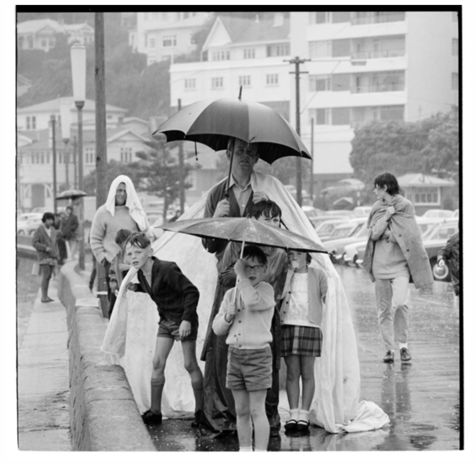 Groups of people sheltering under coats and umbrellas on a wet day at Oriental Bay. Three kids huddle under their dad's rain cape.