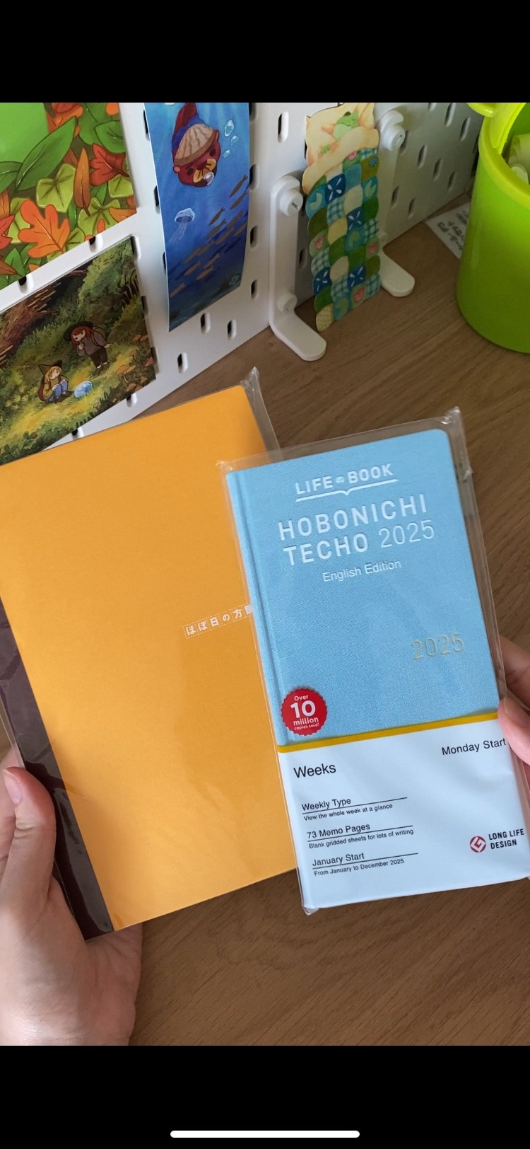 Pic of two hands holding up a yellow hobonichi graph paper a5 notebook and a blue hobonichi weeks that’s still wrapped up for 2025