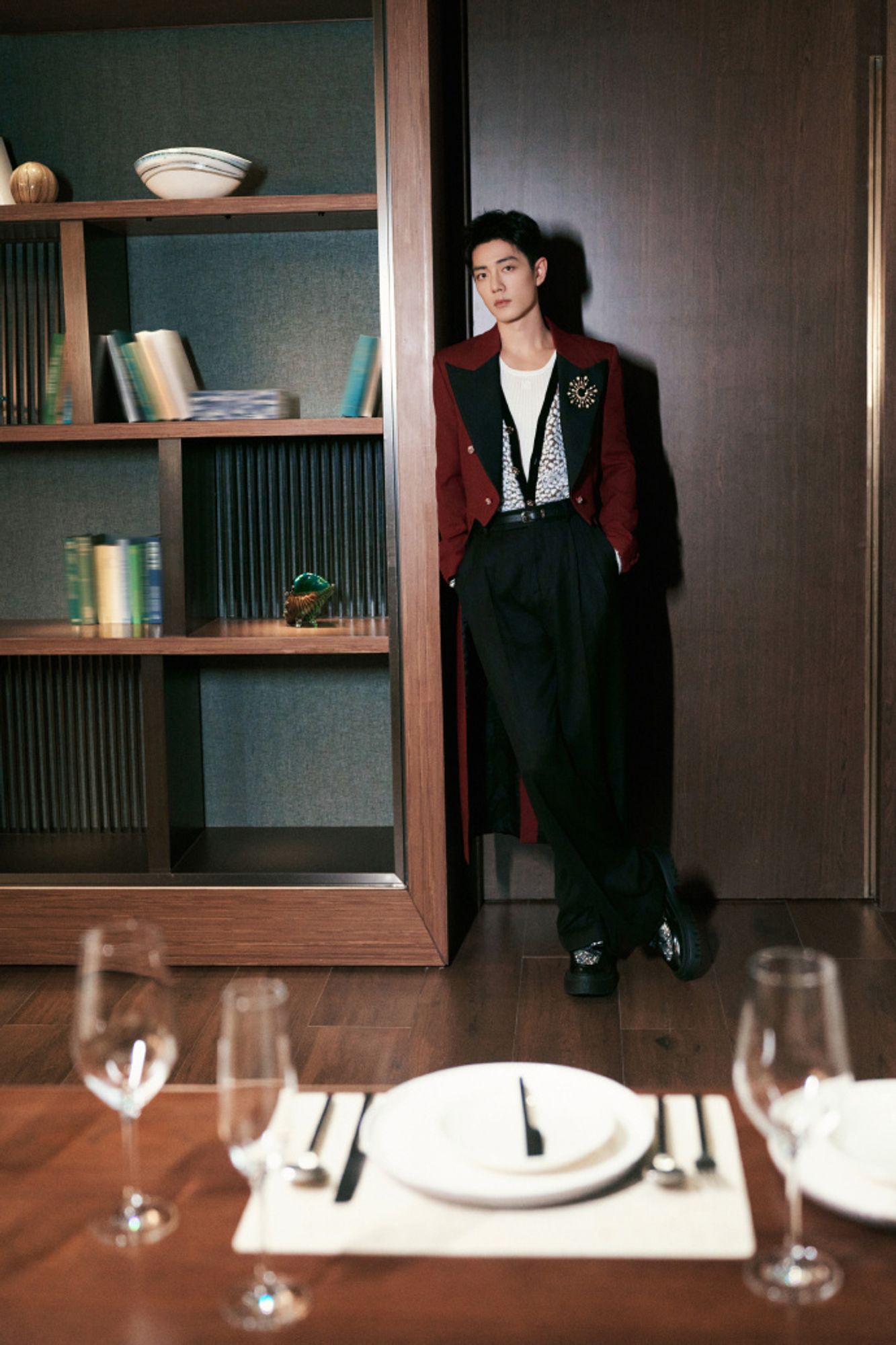 Chinese actor Xiao Zhan, in an elegant room, with dark wood floors and matching bookcases featuring scant books and some objets d'art, blurred in the foreground is fancy place setting. XZ stands further back, leaning against the bookcase, hands in pockets, ankles crossed. He's wearing clunky loafers with large metal fastenings, black dress pants, a white tank, a sparkly silver vest with black edging unbuttoned in deep v, and long red tailcoat with wide black lapels featuring a wide gold brooch. He's looking directly at the camera, expression almost wary.