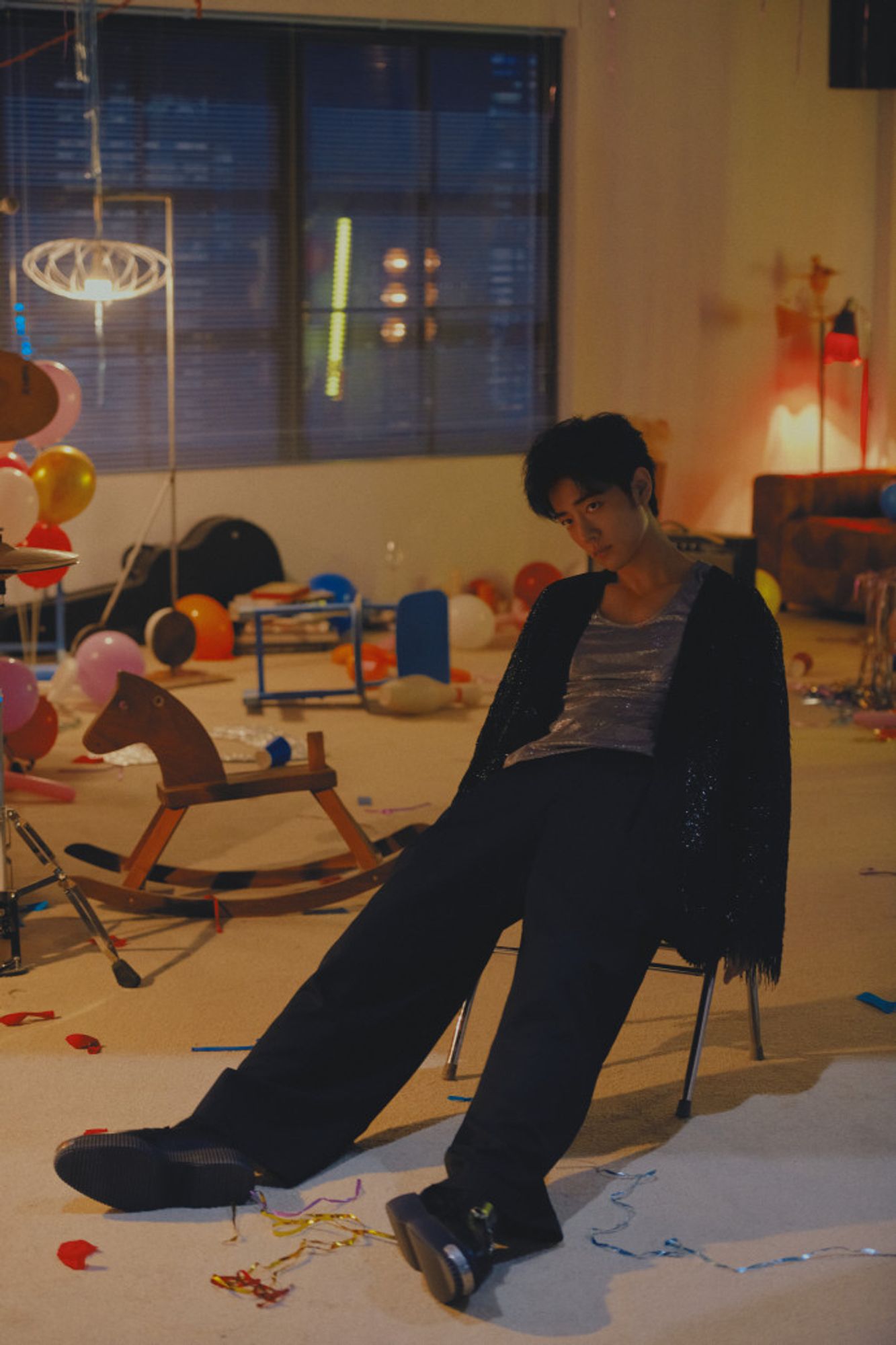 Chinese actor and singer, Xiao Zhan in light colored room littered with party debris (balloons, a ricking horse, streamers, all a mess on the floor). He sit slumped down, legs out stretched in the foreground, wearing black pants, a sparkly silver tank, and a glittery fringed black jacket. His expression is the blankness of exhaustion and being totally over something.