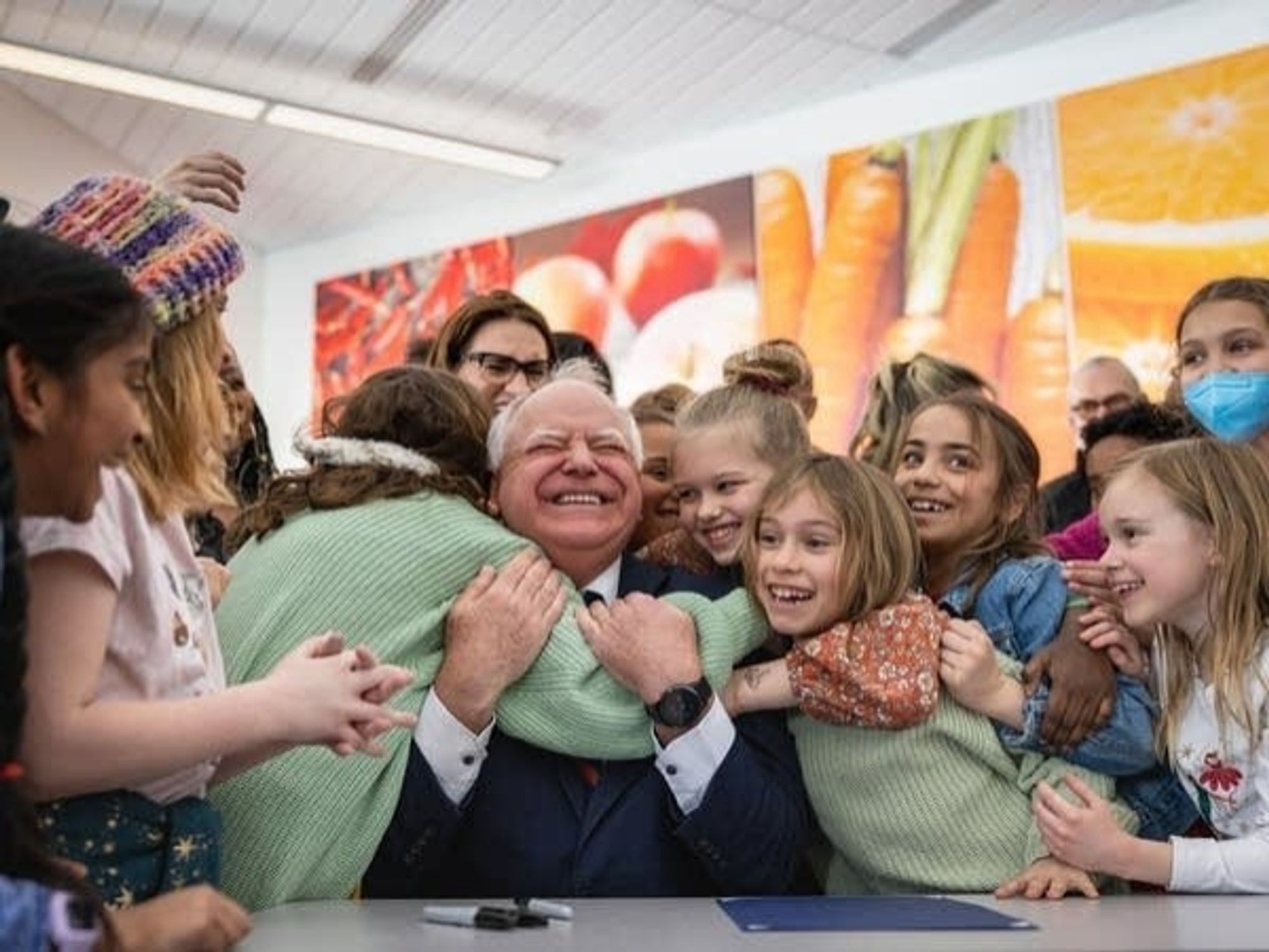 A buncha kids hugging Tim Walz because he gave them free lunch. He has a big smile. Pretty cute.