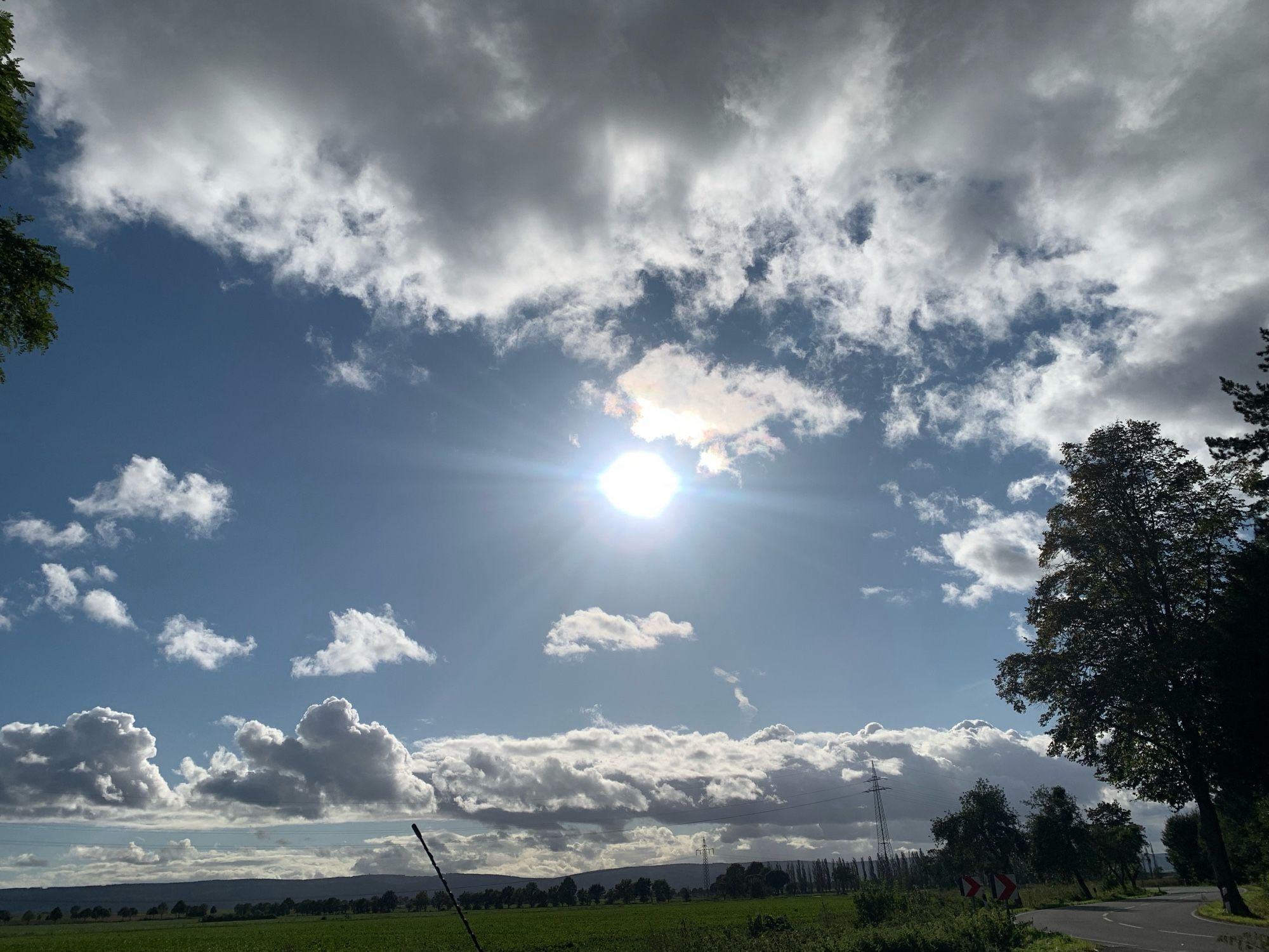 Die Sonne heller strahlender Ball bei hellblauem Himmel und weißen Wolke . Am rechten Rand des Fotos sind ein Weg und Bäume zu sehen.