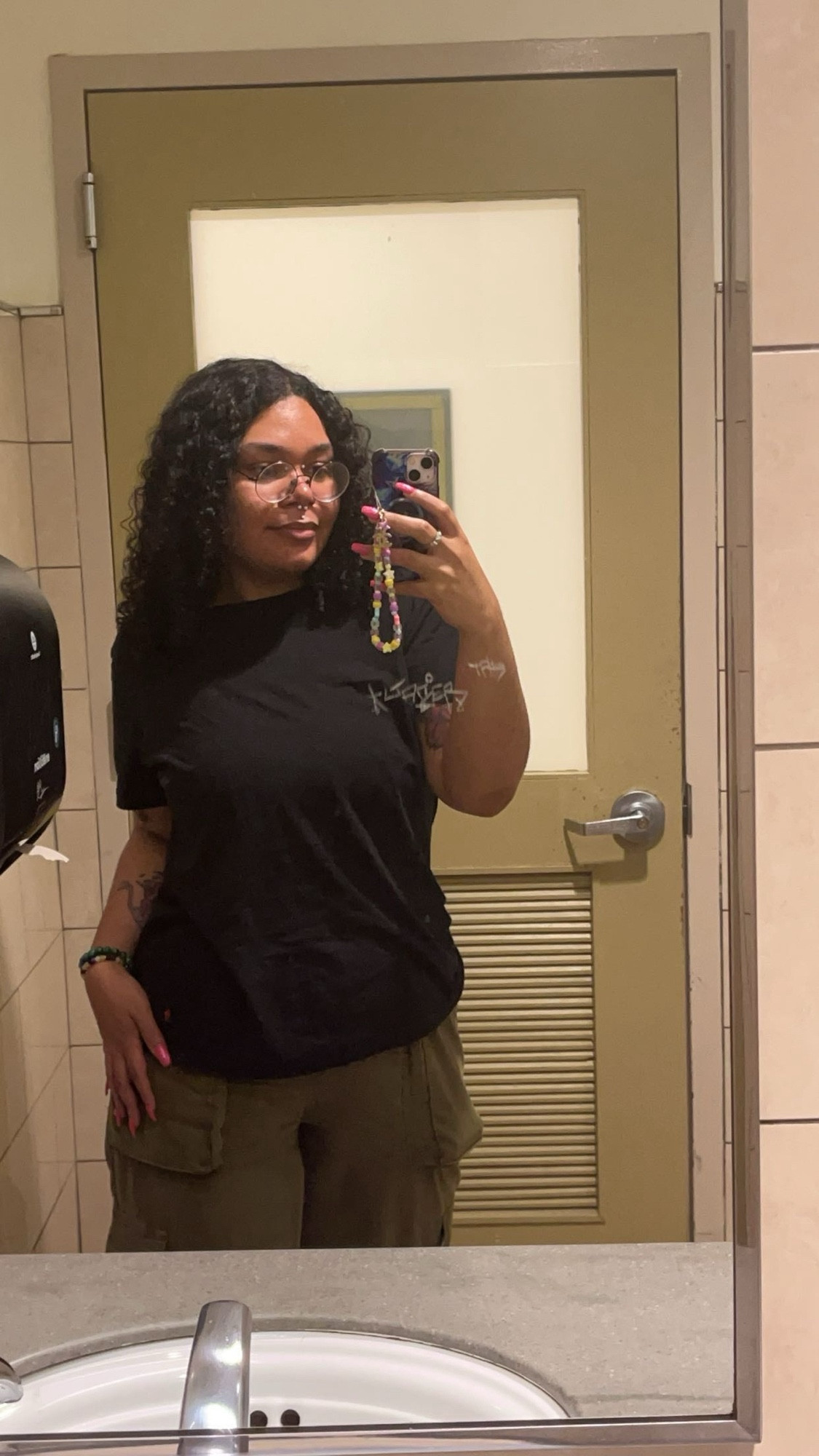 A photo of Lavender standing in a public bathroom, their shirt is plain black and they are wearing avocado cargo pants. One hand is holding their phone and the other is on their hip.