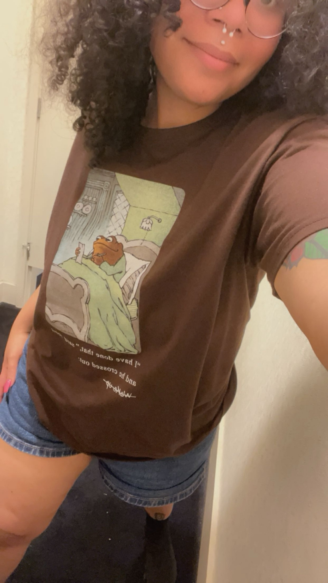 A photo of Lavender in a fitting room wearing blue jeans and a brown shirt with a panel from the book Frog and Toad. Toad is sitting in his bed of his room holding a note pad and pencil.