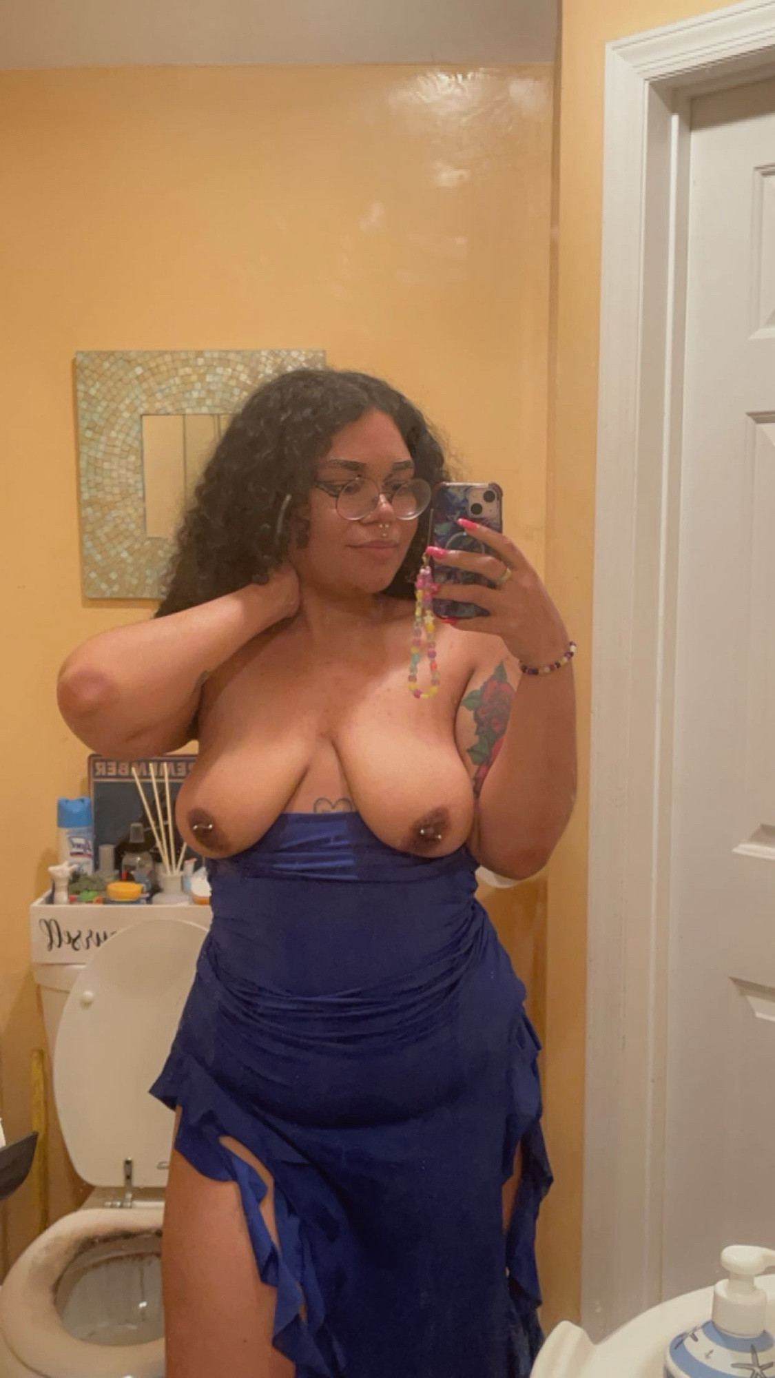 A photo of Lavender standing in their bathroom but the blue tube top dress is lowered revealing their breasts and pierced nipples. Their hand is on their neck and the other is holding their phone