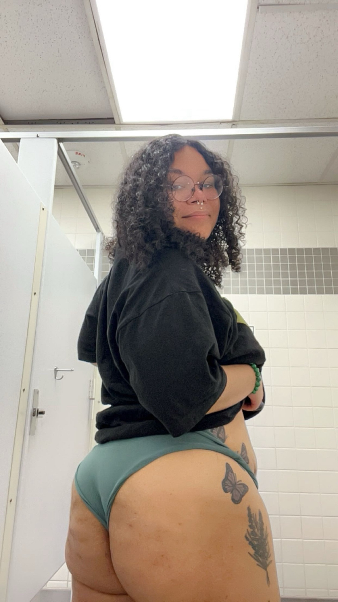 A photo of Lavender standing in a bathroom wearing dark green underwear, their shirt is black and there’s a purple butterfly tattoo on their hip