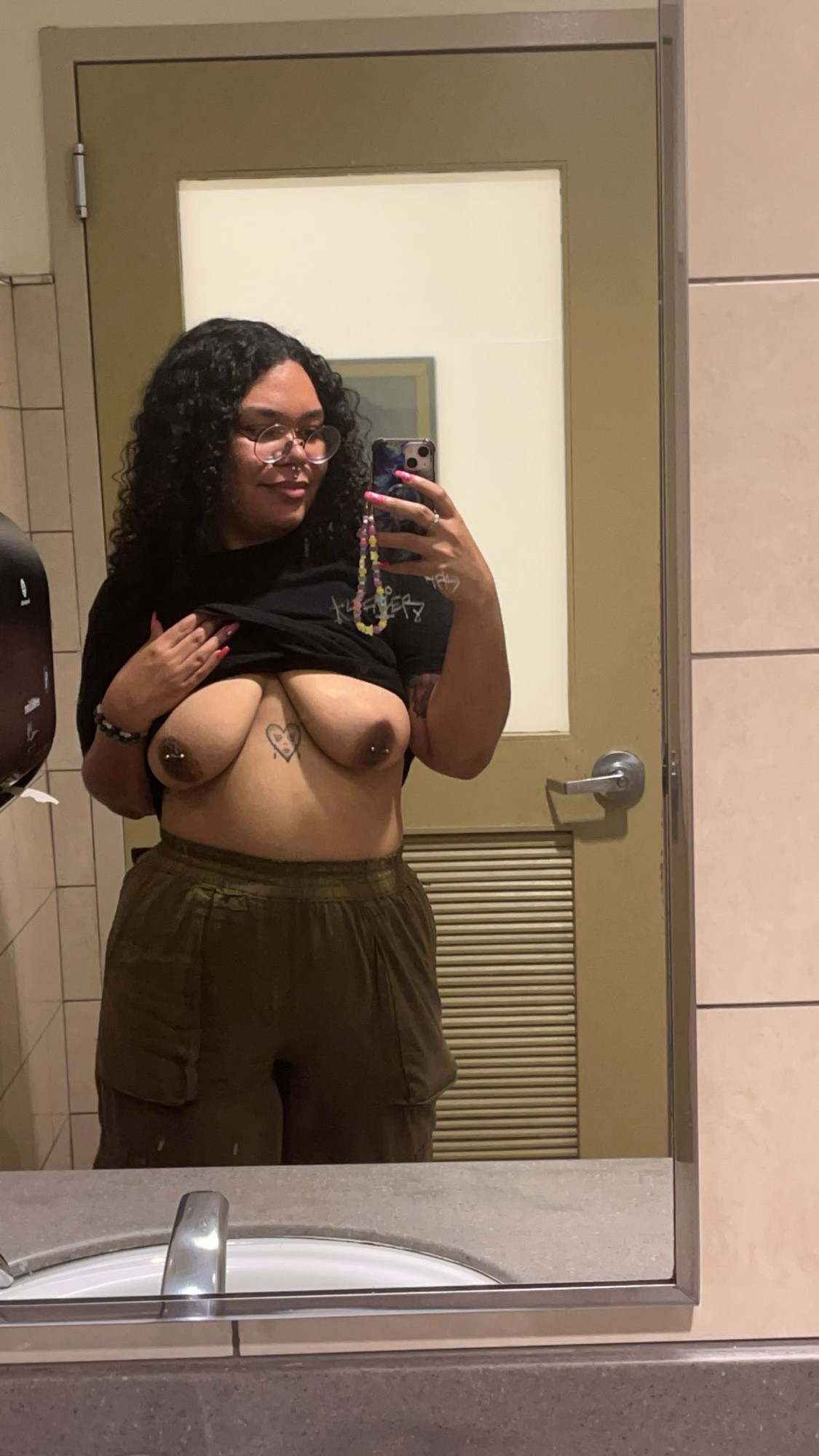 A photo of Lavender standing in a public bathroom, their shirt is plain black and they are wearing avocado cargo pants. One of their hands is holding their phone, and the other lifting up their shirt revealing their breasts and pierced nipples.