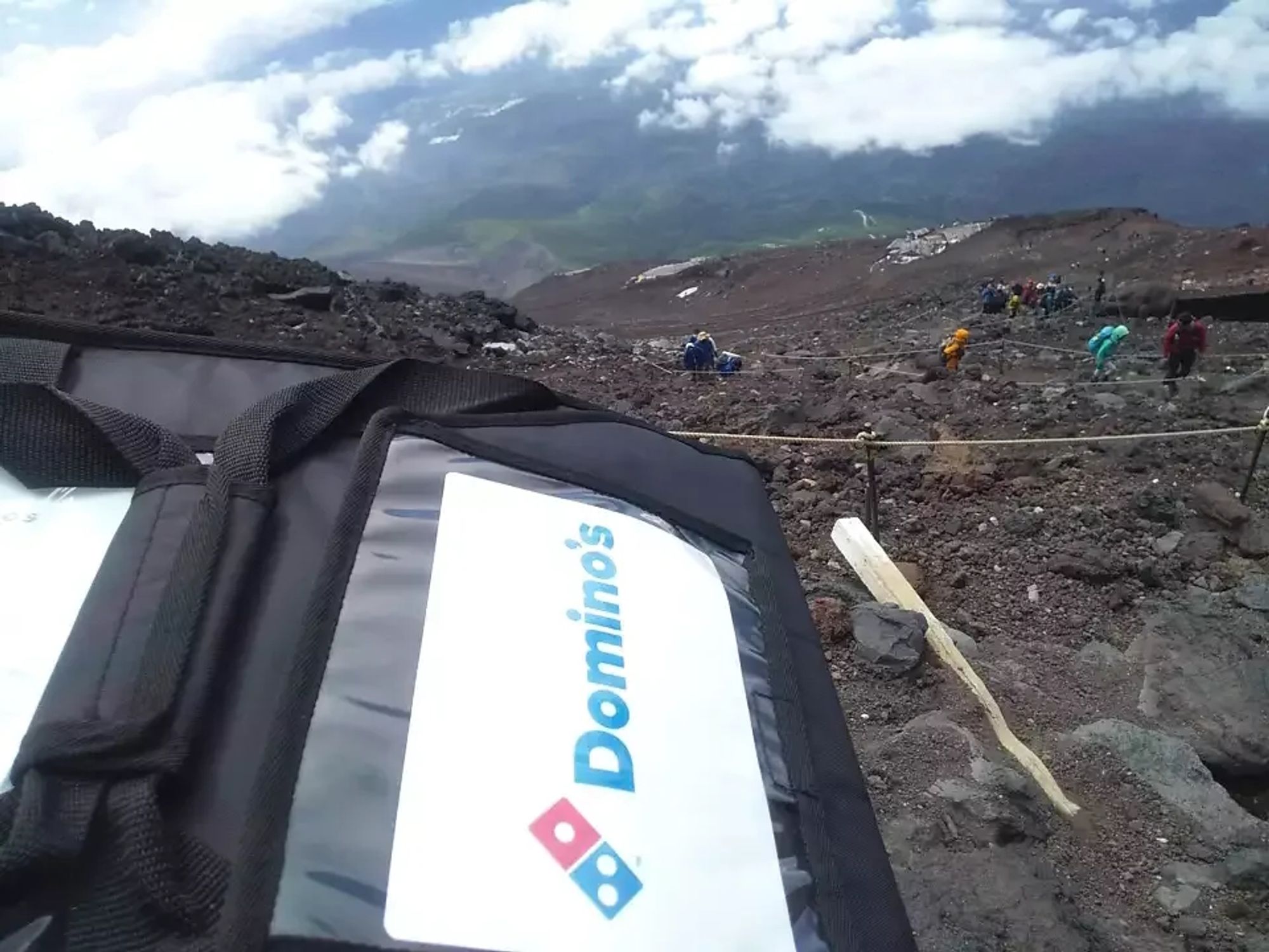 Bild von der Kamera des Pizzaboten über dessen Rucksack in die Tiefe des Berghangs hinab. Man erkennt einen steilen, serpentinenartigen Weg mit Markierungen, auf denen sich vereinzelt Baergwanderer befinden. Er befindet sich über den Wolken, die etwas tiefer im Tal hängen