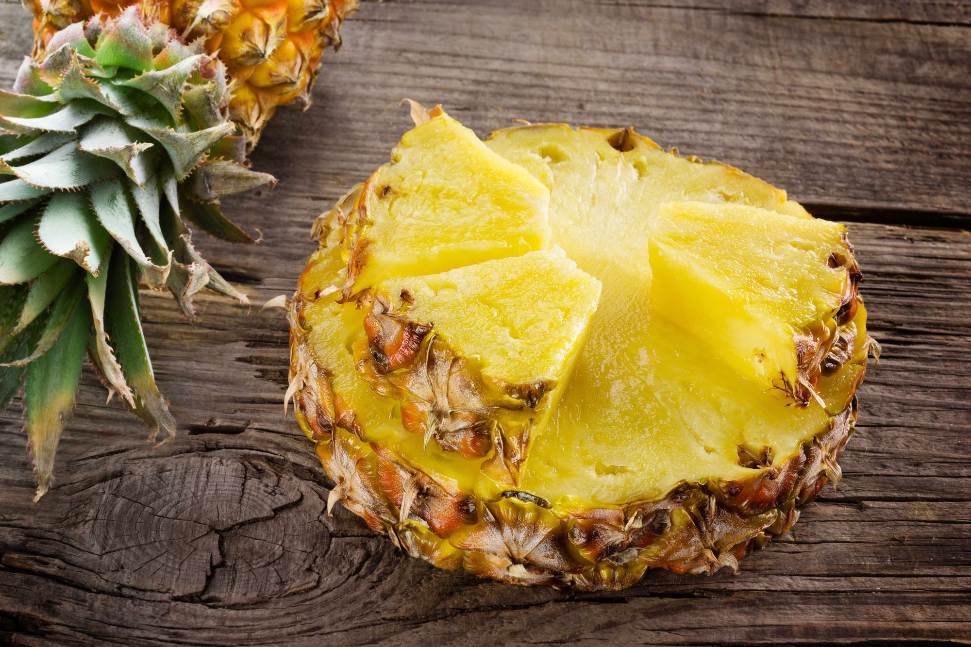 eine halbe Ananas auf einem Holztisch. Drei mundgerechte Stücke liegen auf der Schnittfläche.
Der Strunk liegt links oben neben der Frucht