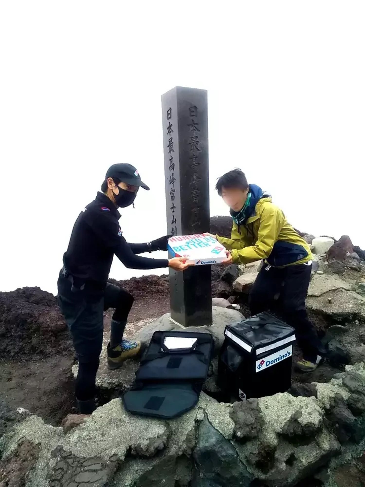 der Pizzabote übergibt seine Lieferung an einen Bergsteiger am Gipfelpfahl (eine steinerne Säule mit Schriftzeichen)