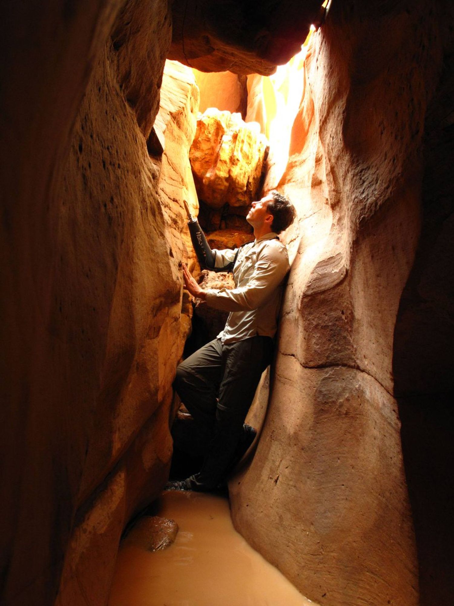 Aron Ralston steht in dem schmalen Felsspalt des Unglückes und schaut nach oben, wo Sonnenlicht sich an den gelbroten Felswänden widerspiegelt. Man sieht, das der Spalt gerade eng genug ist um die Person aufzunehmen, mit leicht angewinkeltem Knie passt er in den Spalt