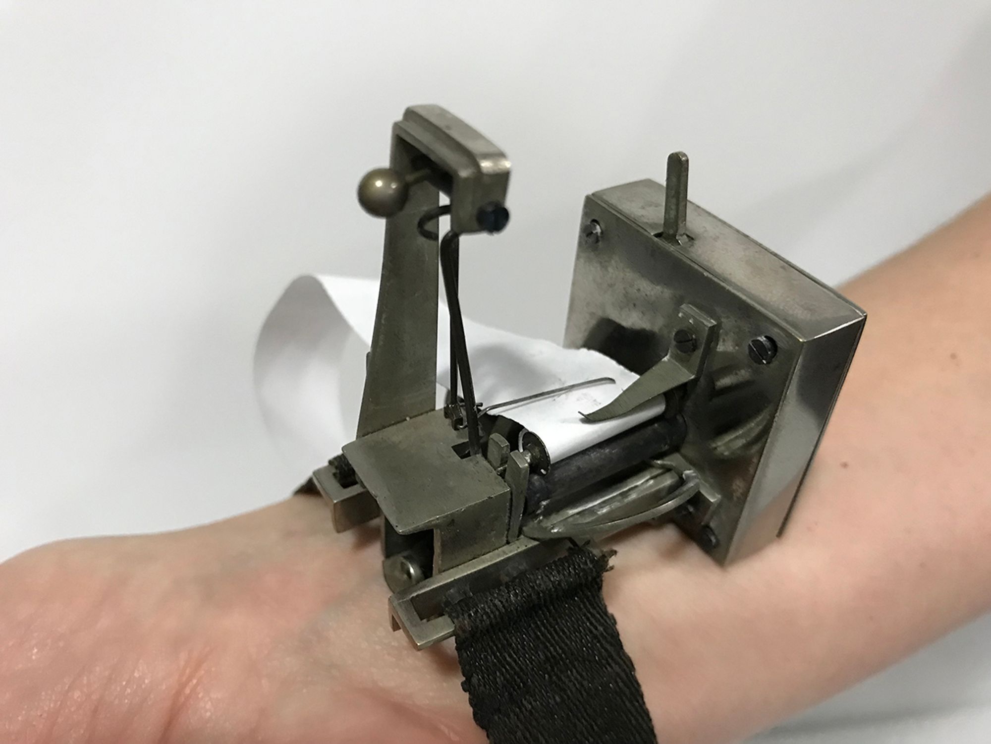 Detailaufnahme eines Sphygmograph am Handgelenk, innen. Ein Kästchen aus Stahl mit diversen Bügeln und Federchen, eine Nadel schreibt auf einen Papierstreifen. Die Apparatur ist mit einem schwarzen Gurt am Handgelenk befestigt