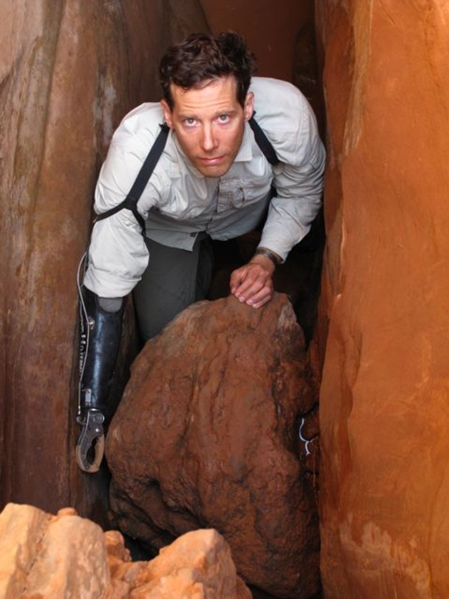 Aron Ralston bei einem späteren Besuch des Unglücksortes. Sein rechter Arm ist ab Ellenbogen eine Prothese. Er steht etwas gezwängt in dem schmalen Spalt und schaut über zwei Felsbrocken nach oben.