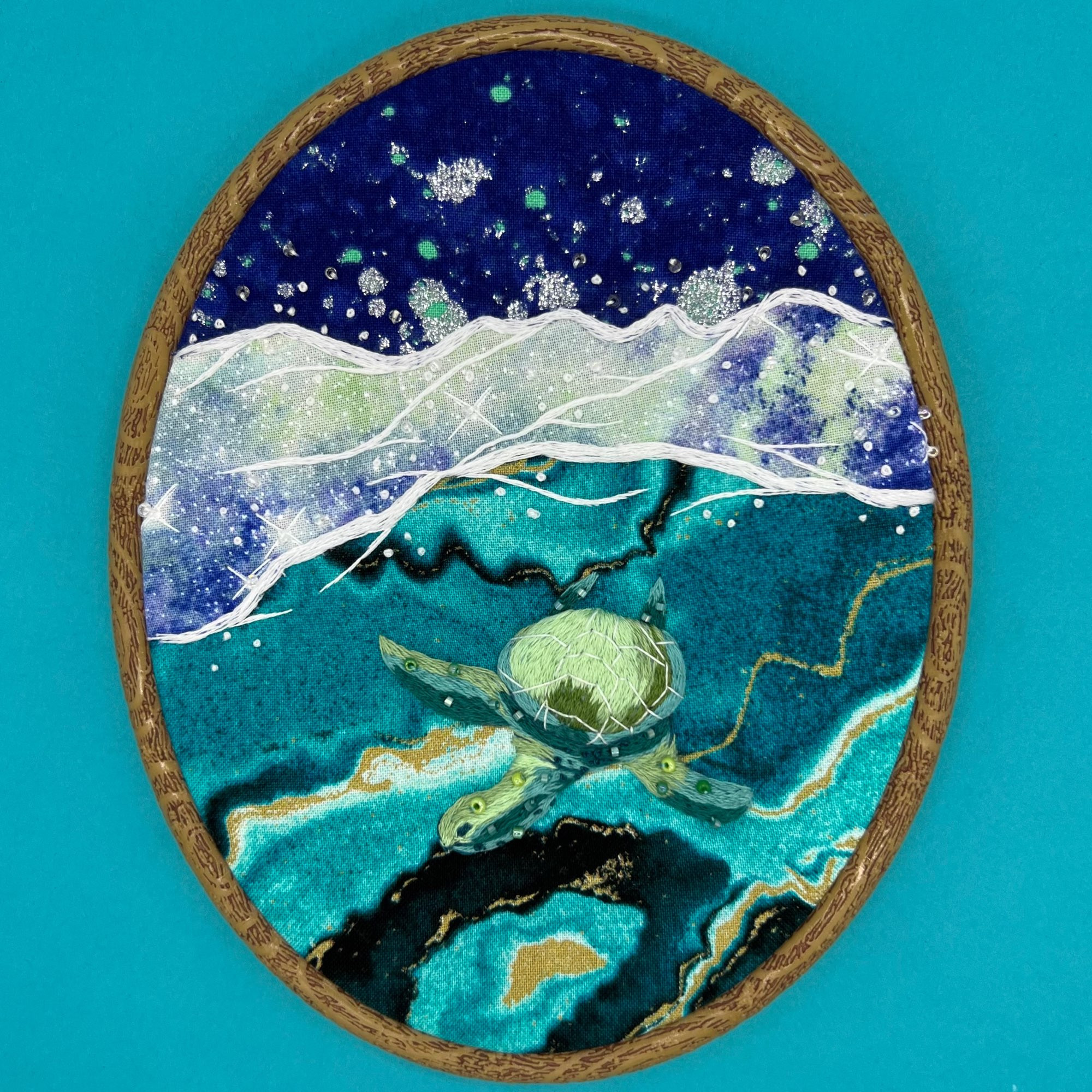 An hand embroidered hoop of a green sea 
turtle on a teal background. The turtle and sea foam is stitched with thread and a small about of beads over a collage of fabric mimicking water and stars in various shades of blue and green.