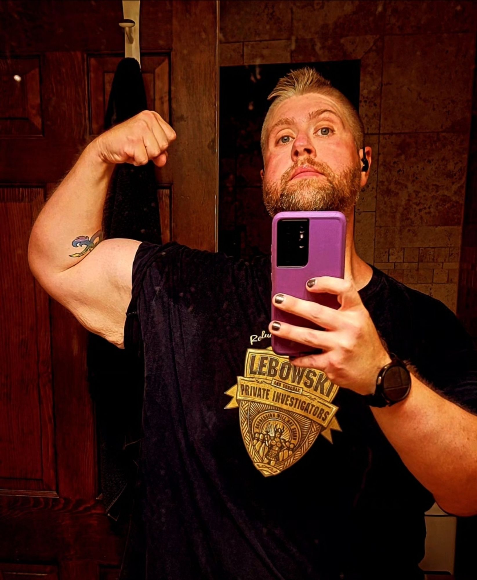 The image shows a bearded man posing in front of a bathroom mirror, flexing his arm to showcase his bicep. He has a light blonde, short, slightly spiked hairstyle, and is wearing a dark blue shirt with a graphic that reads "Lebowski Private Investigators." His arm features a visible tattoos, and he has painted nails, adding a bold and expressive touch to his look. He holds a purple phone to capture the selfie with a confident expression.